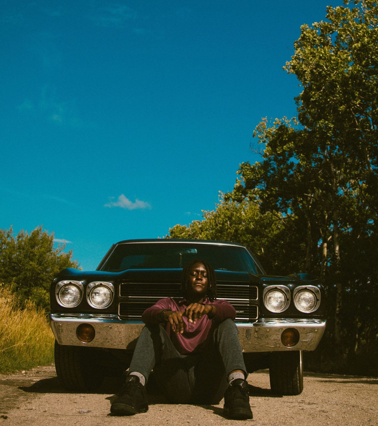 A young man sits relaxed in front of a classic black muscle car on a dirt road surrounded by green trees under a blue sky.