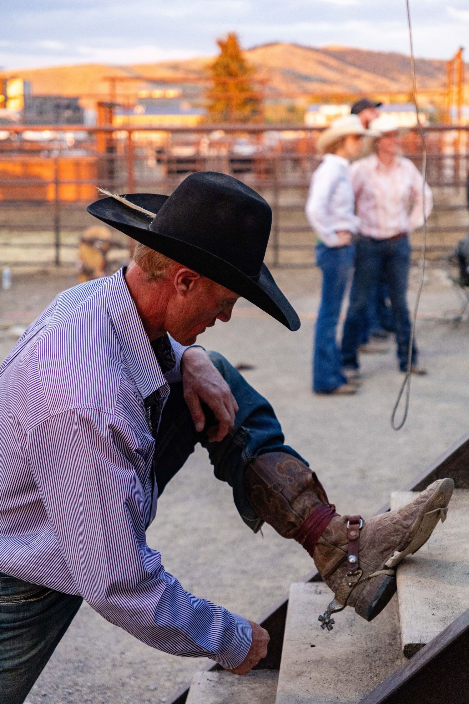 Cowboy in black hat adjusts boot spur before rodeo competition with mountains glowing at sunset.