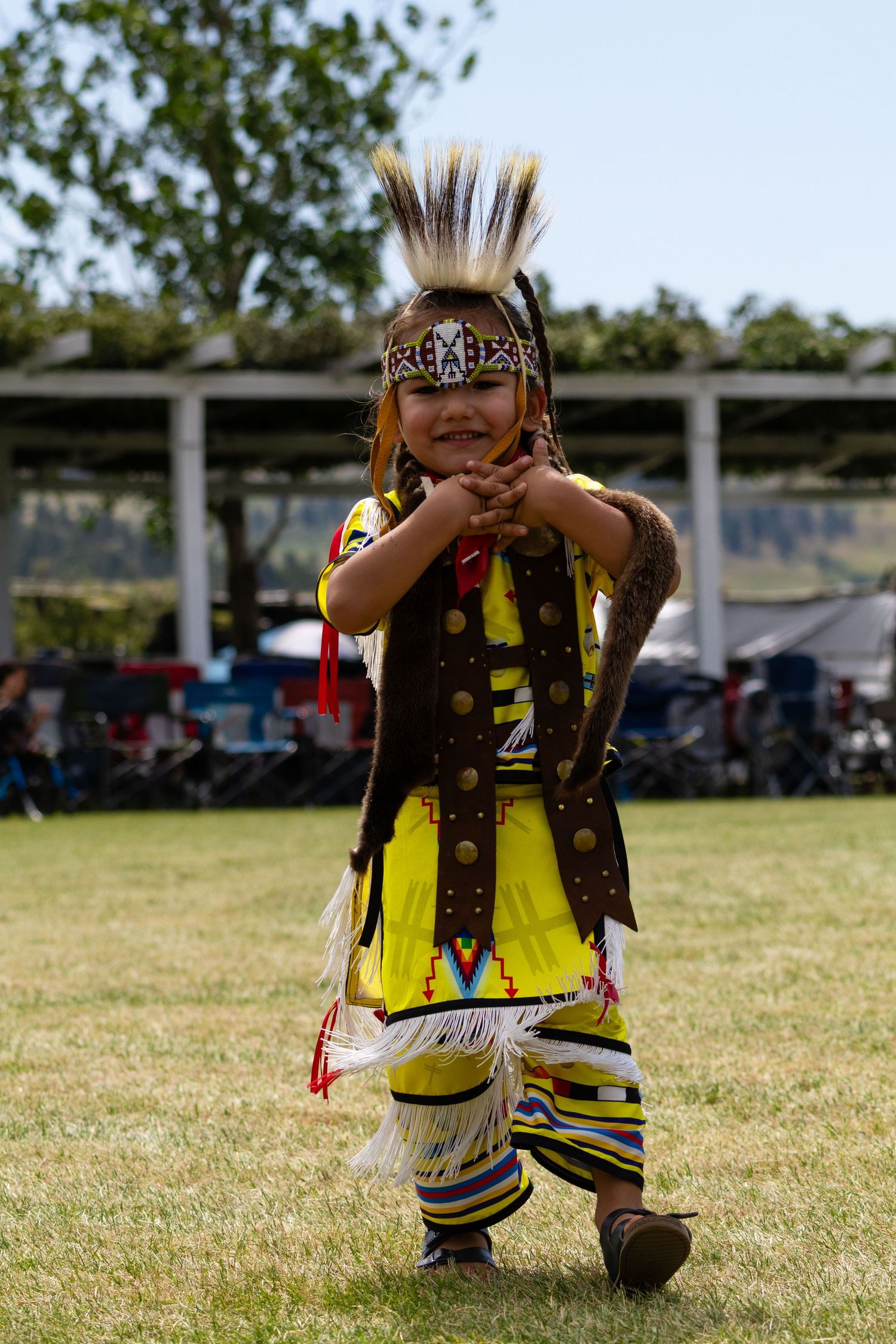 Young Native American boy in yellow fringed regalia and feathered headdress dancing at powwow.