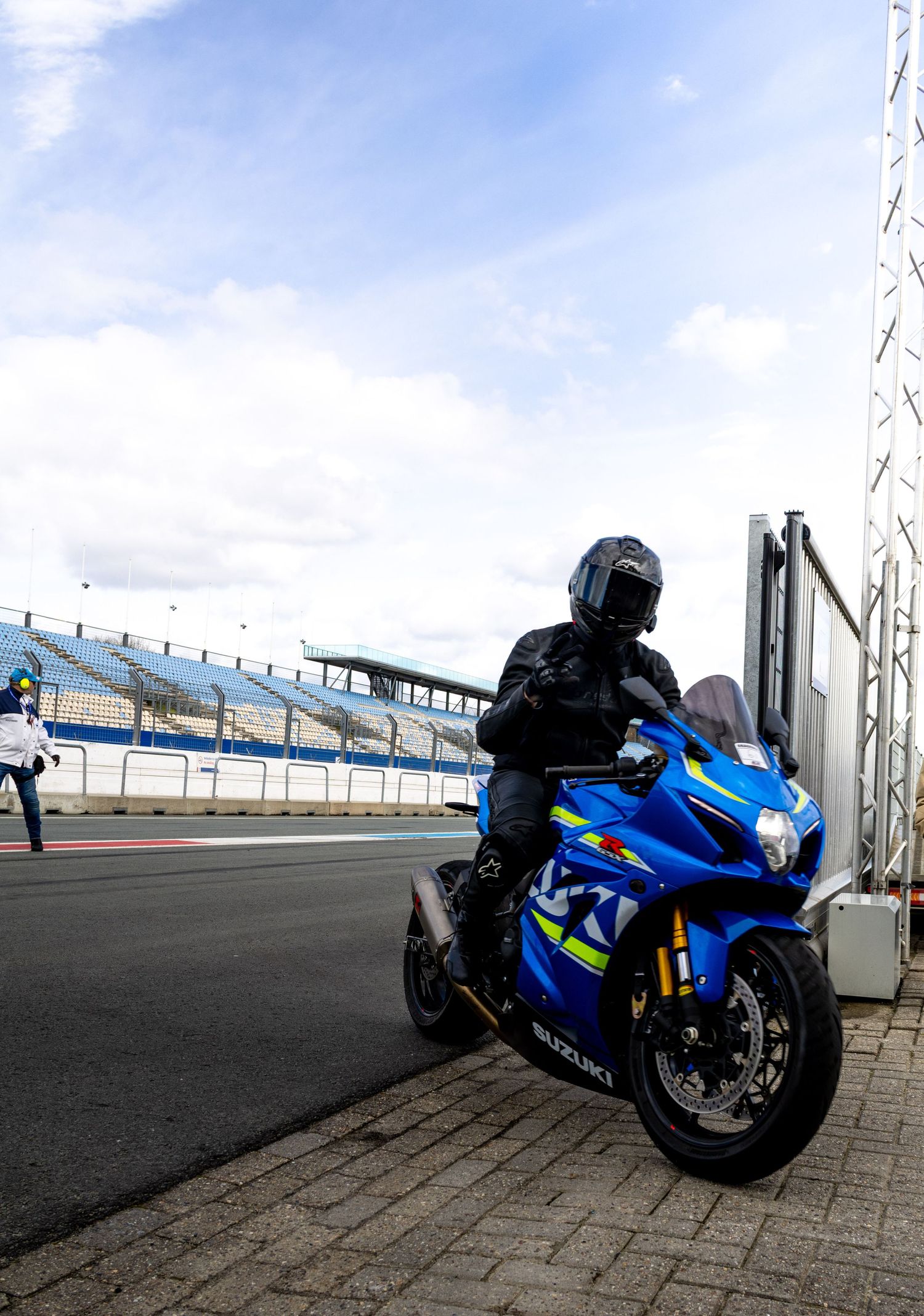 Rider in black gear sits on a blue Suzuki GSX-R motorcycle at a racing circuit with grandstands in the background.
