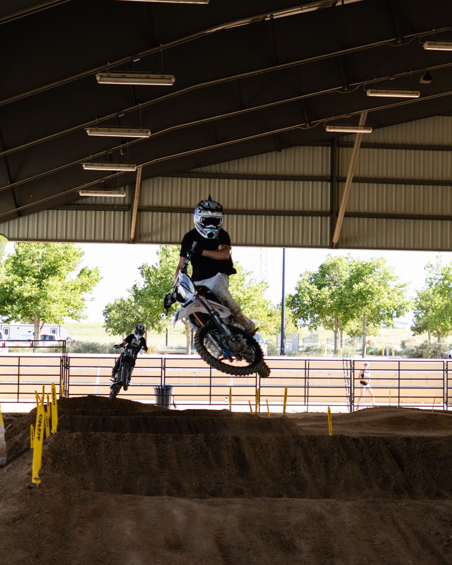 Dirt bike rider jumps over tabletop obstacle at indoor motocross track with second rider behind.