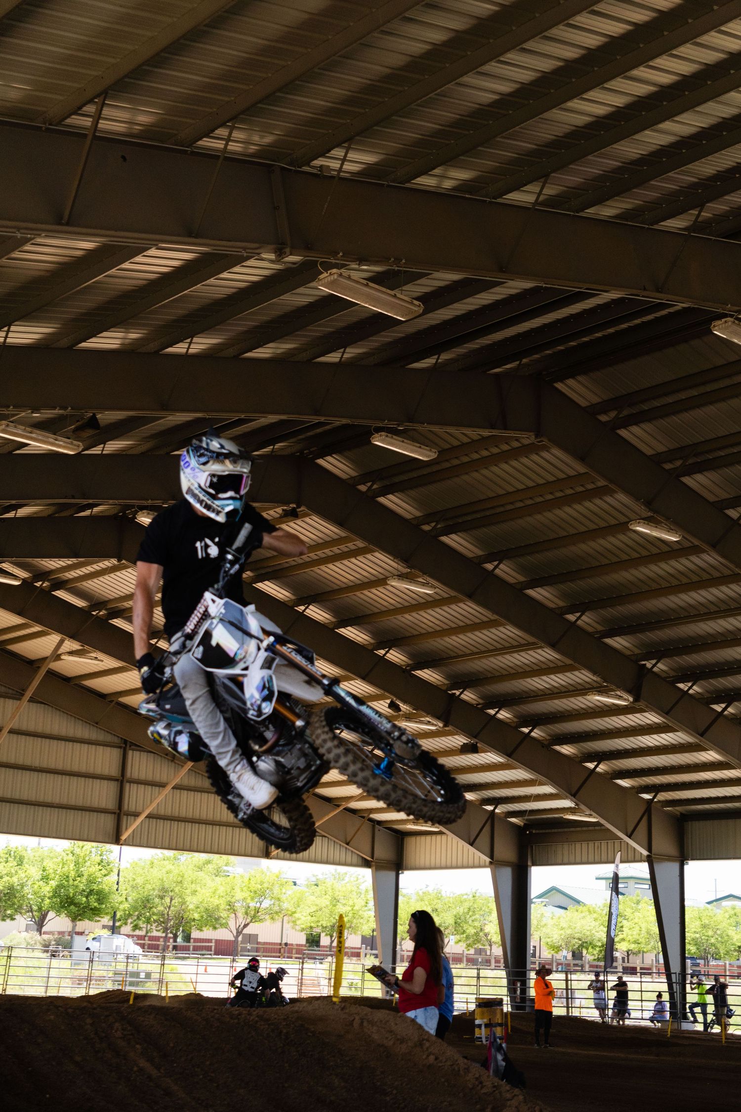 Motocross rider in white gear soars over a dirt jump inside a large covered indoor supercross arena.