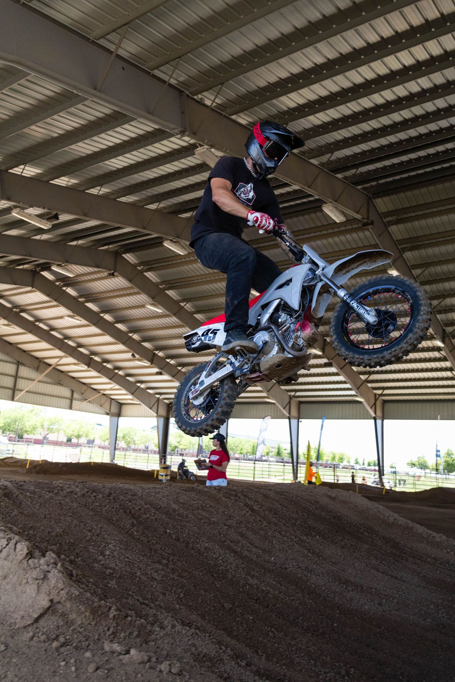Motocross rider catching air on a jump inside a covered indoor dirt track during a race event.