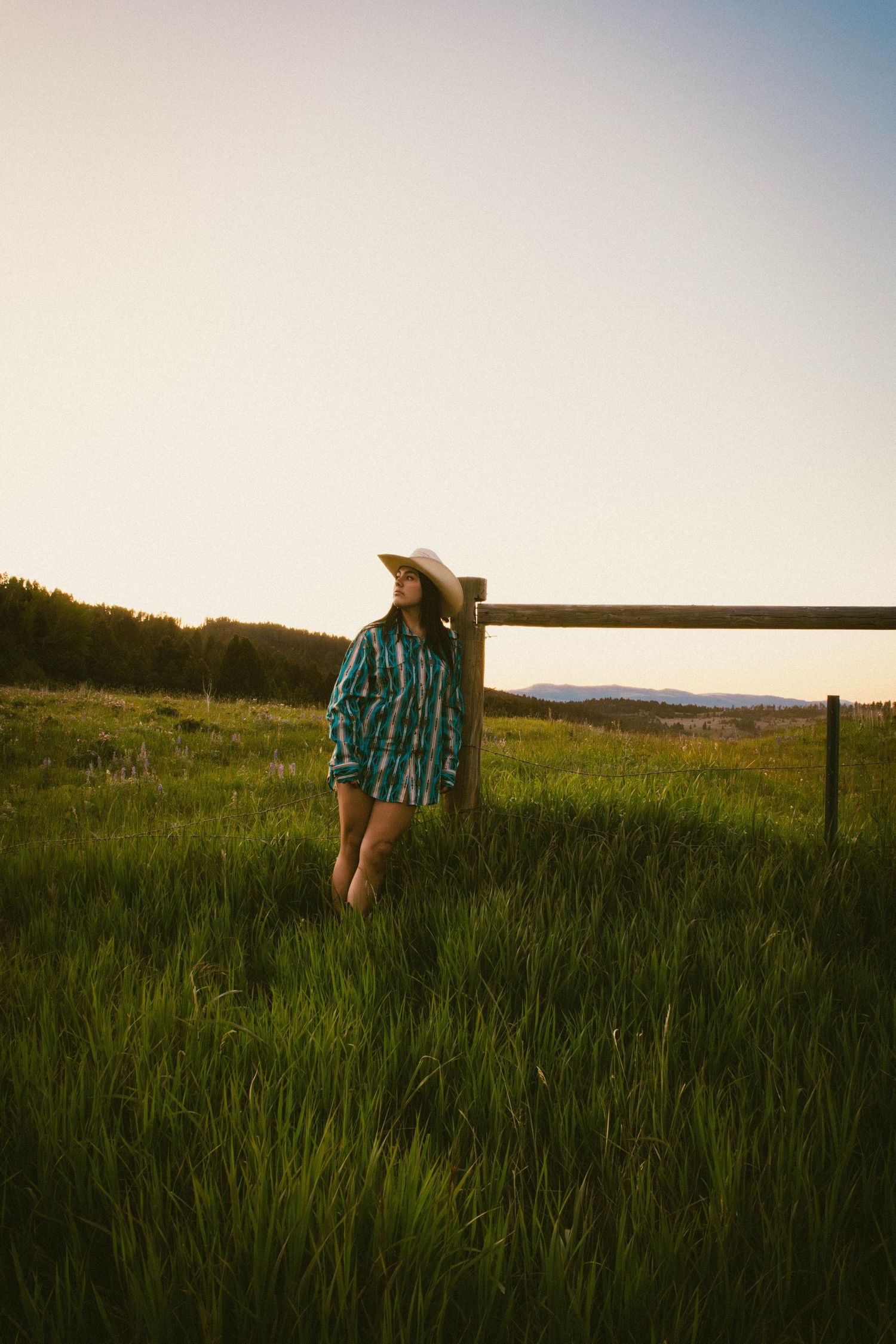 Woman in teal plaid shirt and white cowboy hat leaning on a wooden fence post in a green meadow at sunset.