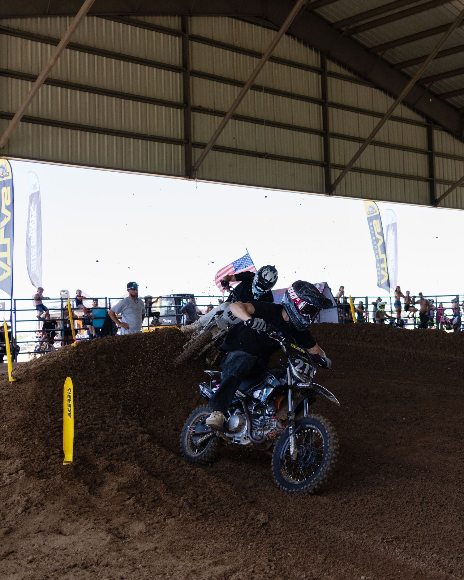 Motocross rider carries American flag while navigating dirt track inside covered arena at competition.