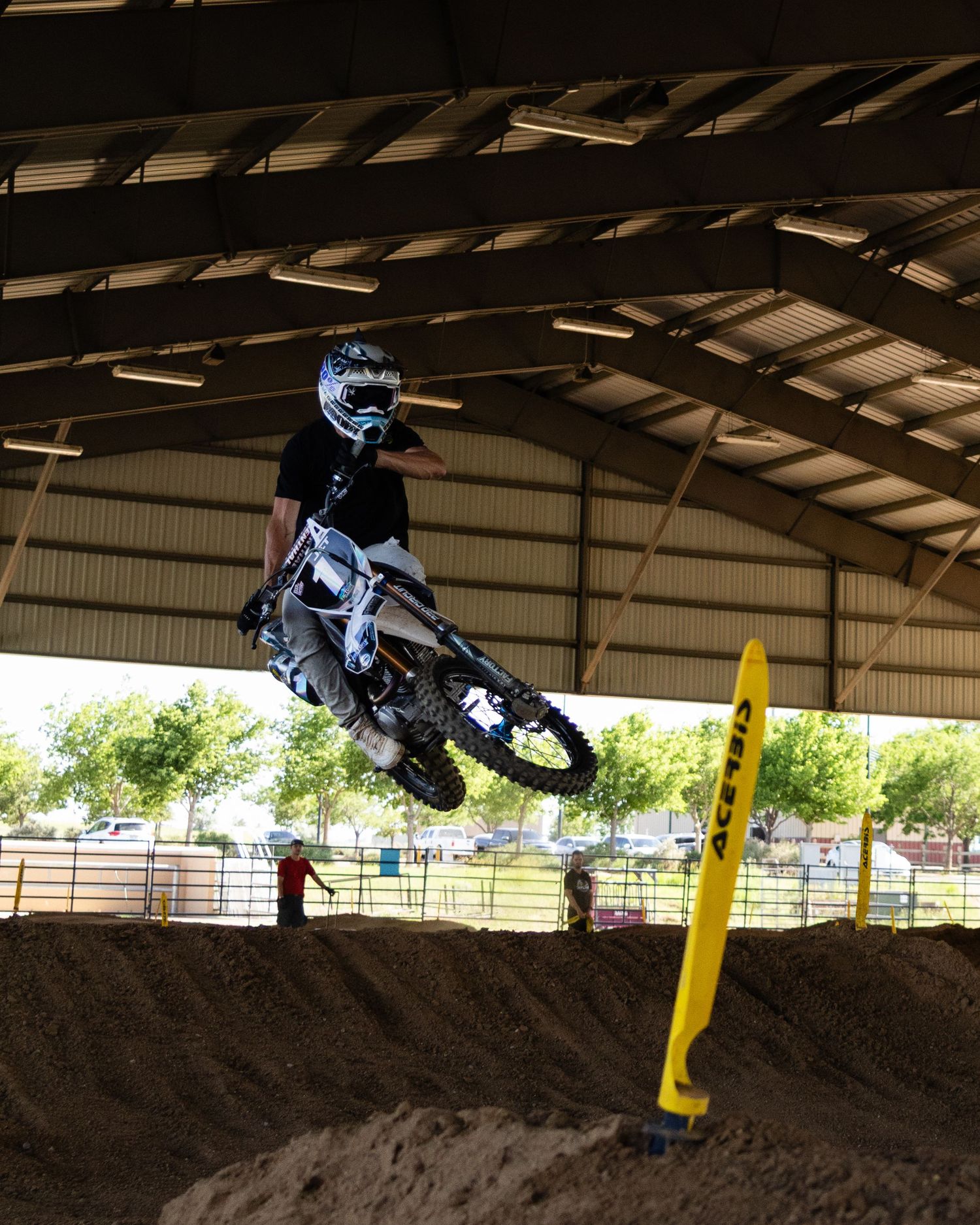 Dirt bike rider performing a jump over a mound at an indoor motocross track.