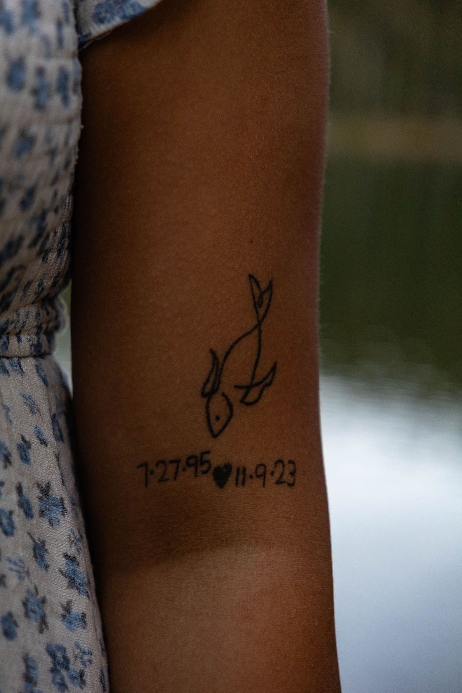 Close-up of an arm tattoo featuring a fish design with dates 7-27-95 and 11-9-23.