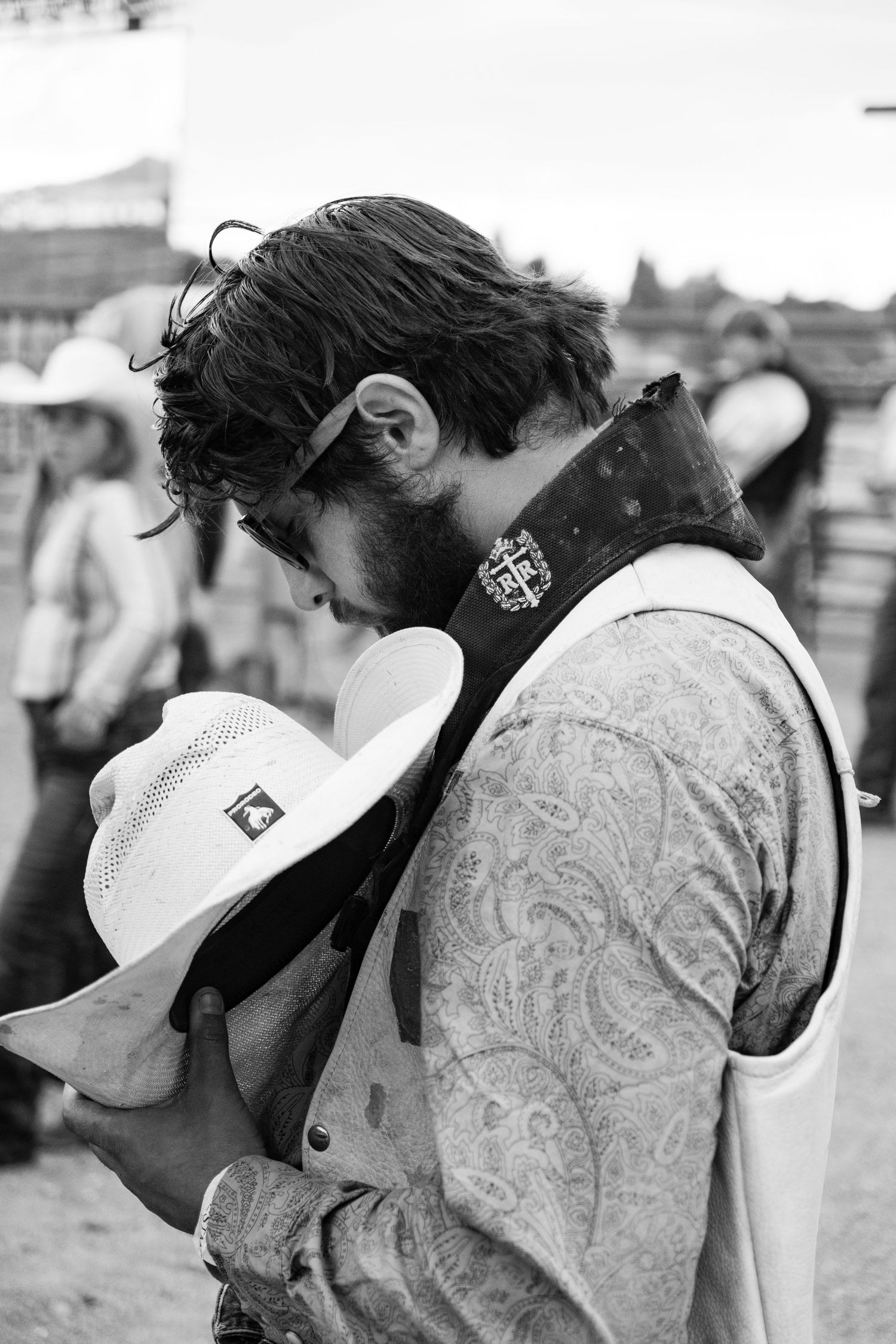 Black and white portrait of bearded cowboy bowing head over white hat at a rodeo event.
