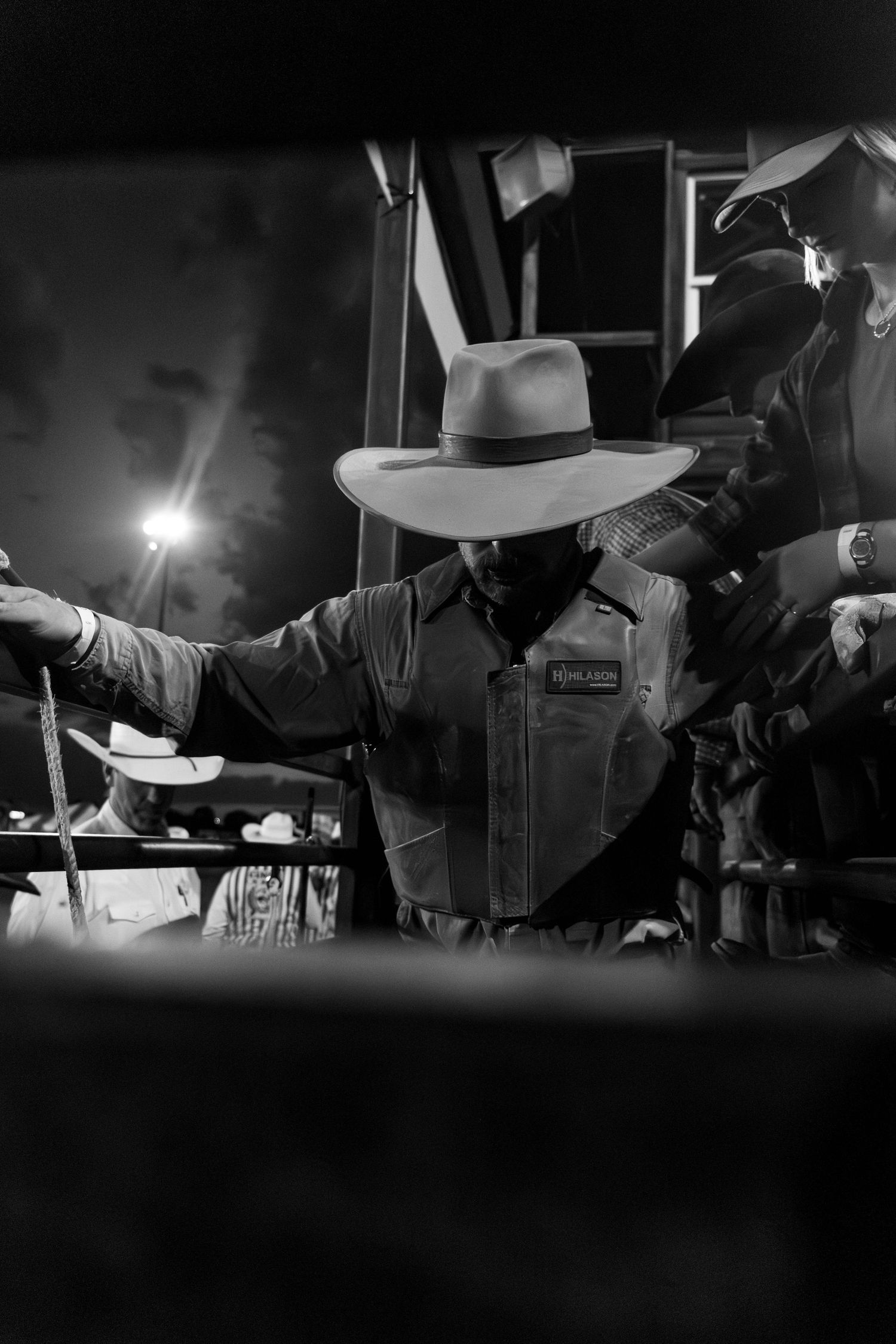 Black and white photo of a cowboy in hat and vest preparing at a rodeo chute at night.