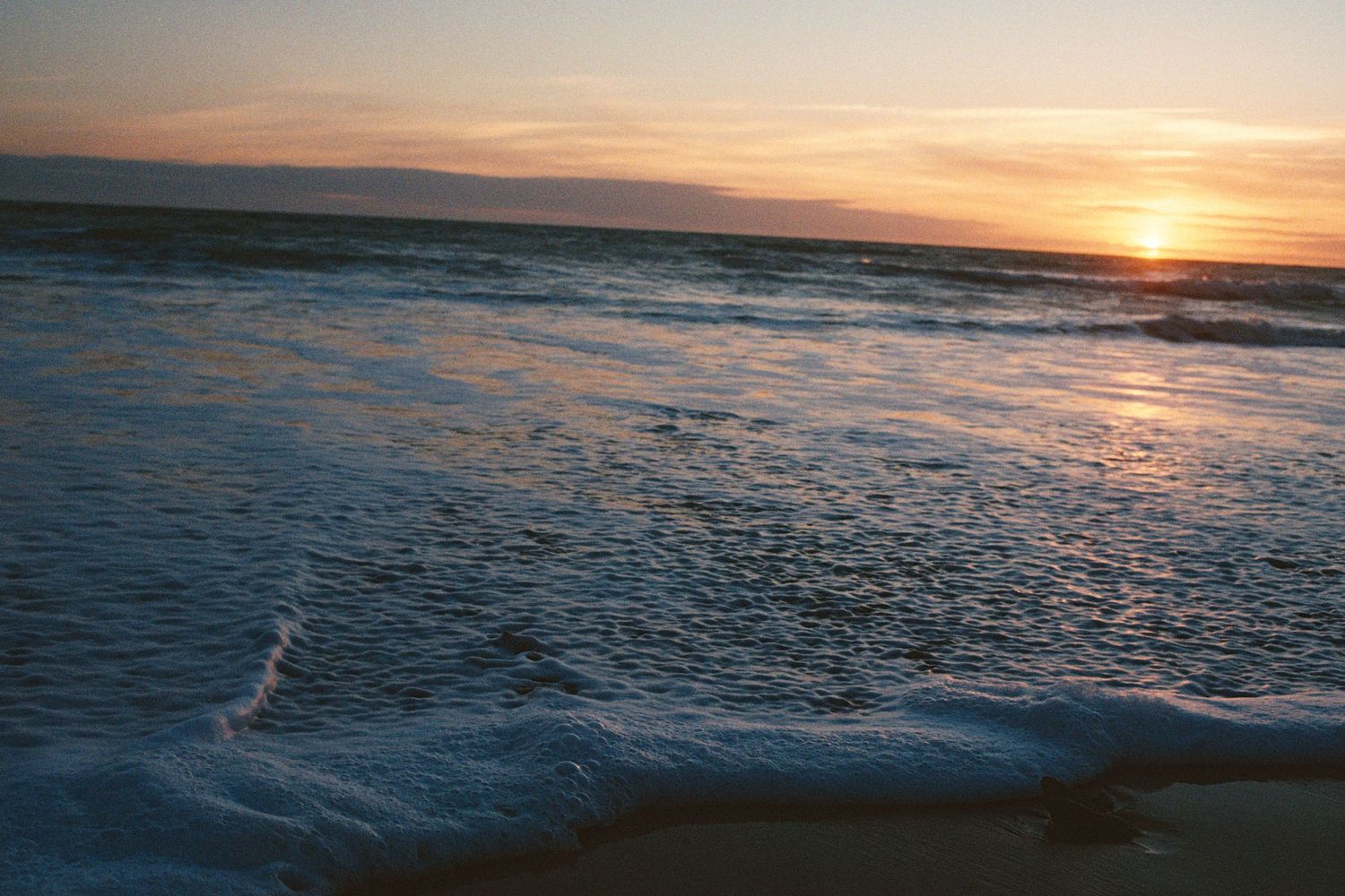 Gentle ocean waves rolling onto sandy beach during a colorful sunset over the horizon.