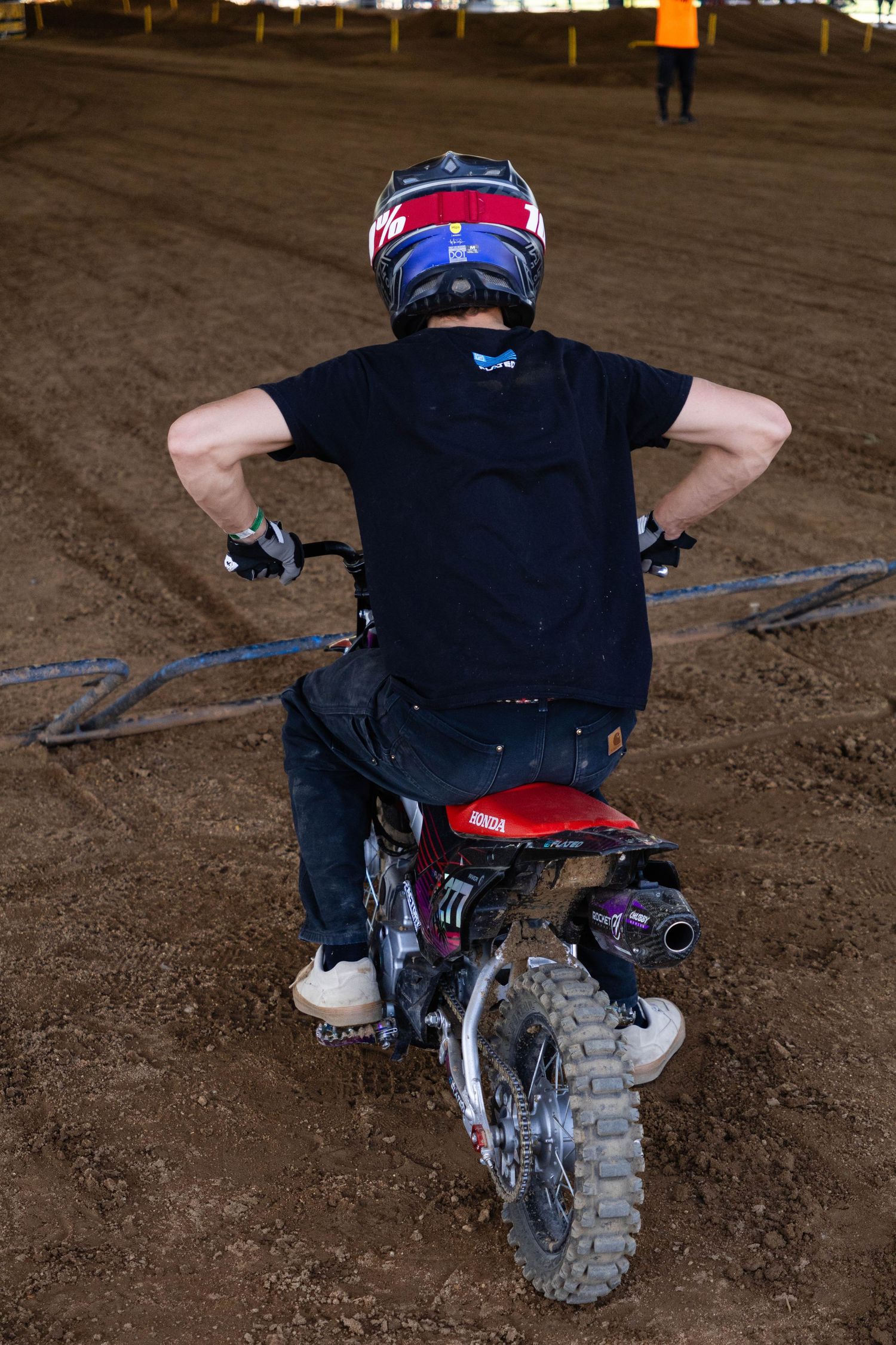 Rear view of a motocross rider on a small red dirt bike wearing a colorful helmet in an indoor arena.