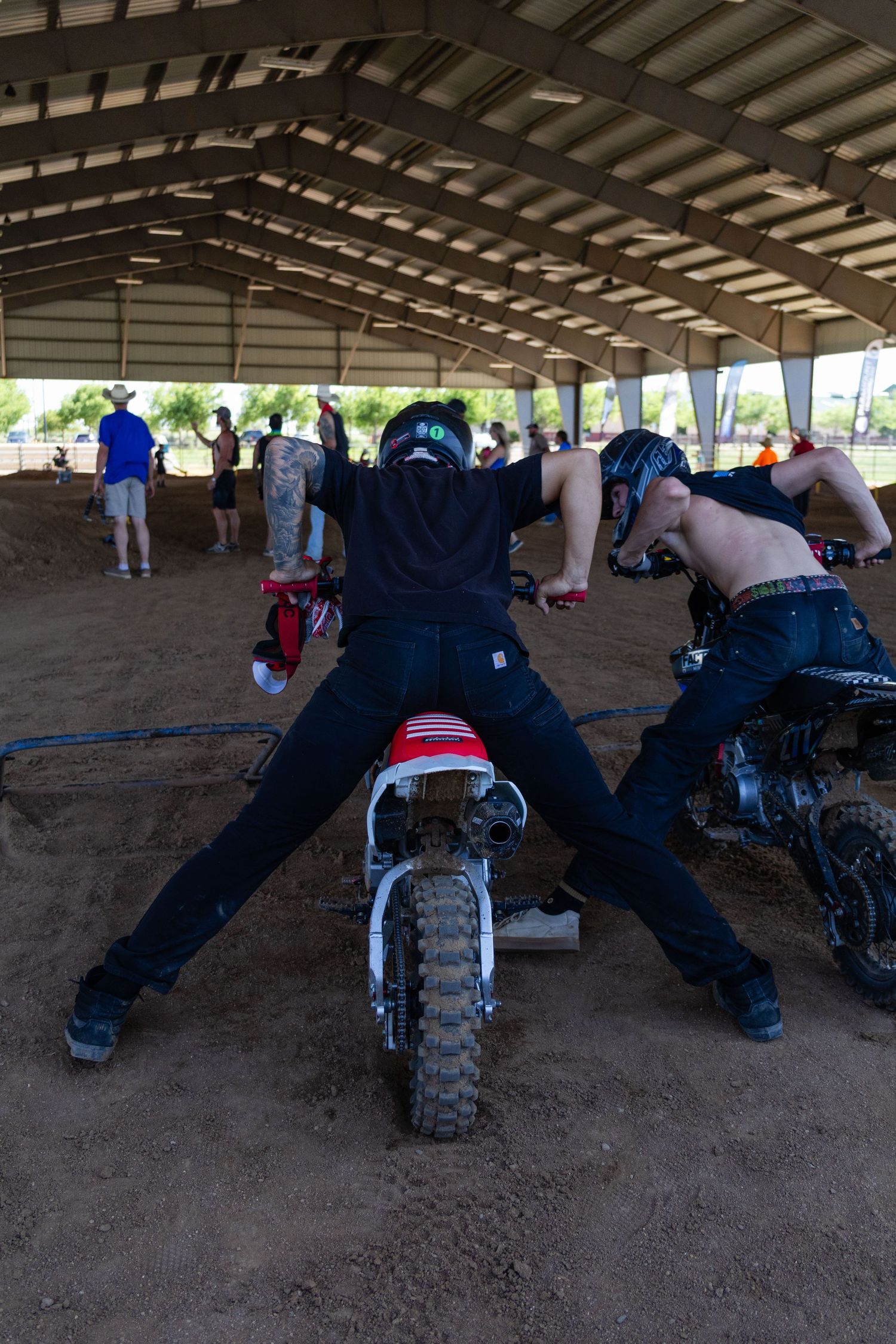 Two motocross riders in helmets straddle dirt bikes from behind inside a covered dirt arena.