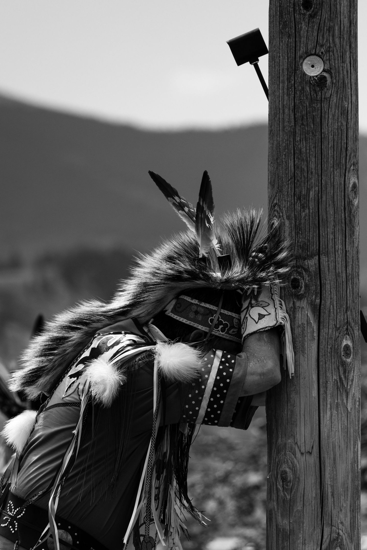 Black and white photo of Native American dancer in fur regalia leaning against wooden pole.