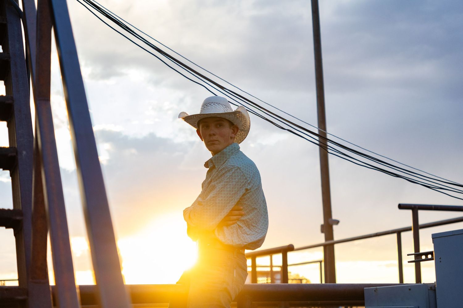 Young cowboy in white hat leans on railing silhouetted against golden sunset at outdoor rodeo.