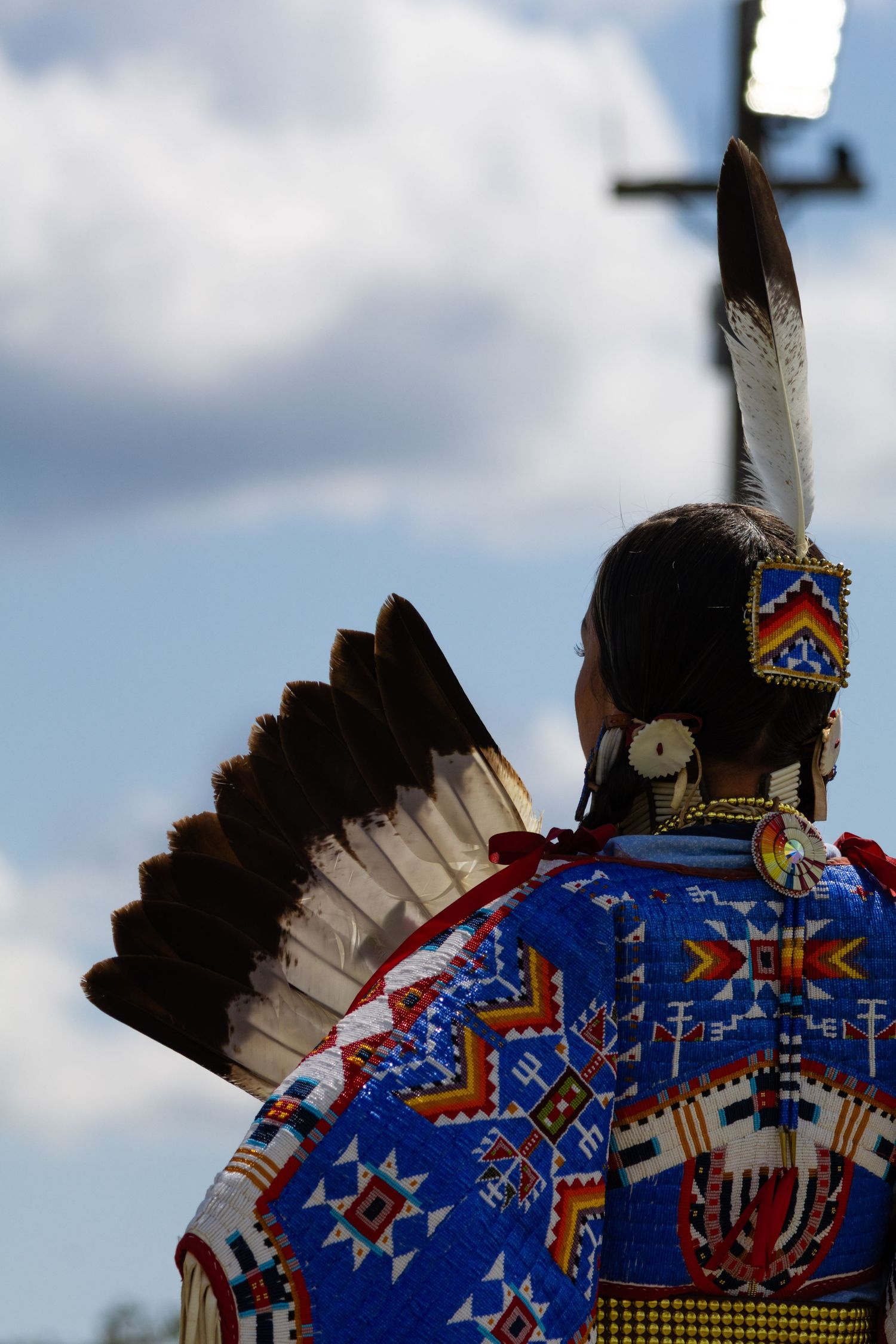 Back view of Native American dancer in blue beaded regalia with feathered fan at an outdoor powwow.