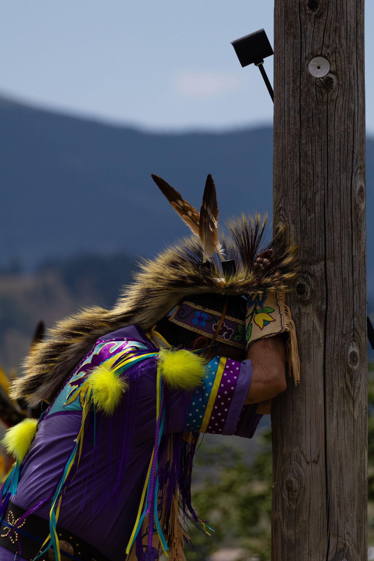 Native American dancer in fur and feathered regalia resting against wooden pole at powwow.
