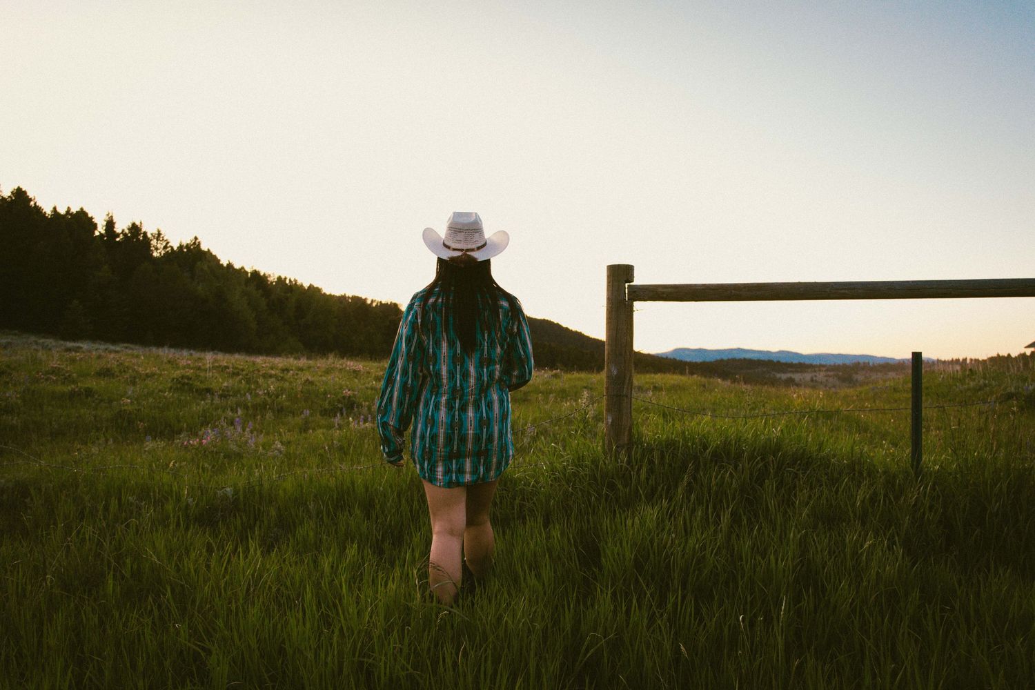 Woman in turquoise plaid shirt and white cowboy hat walking toward a wooden fence at sunset.