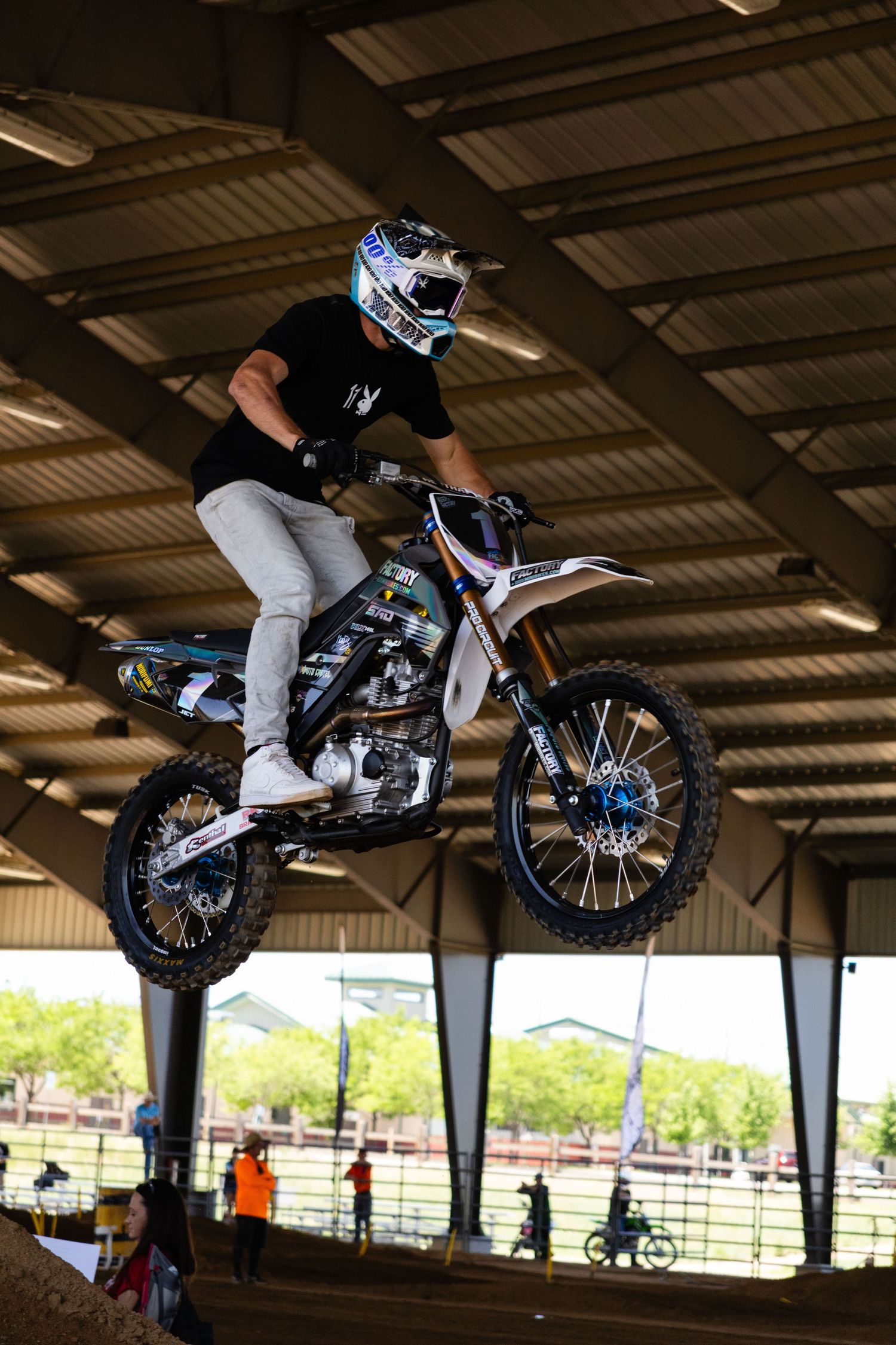 Motocross rider performing a jump on a dirt bike inside a covered arena at a motorsport event.
