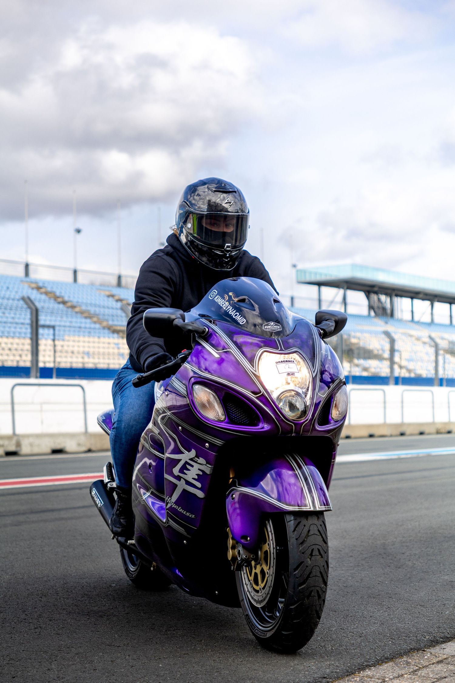 Rider on a purple Suzuki Hayabusa motorcycle on a racing circuit with empty grandstands in background.