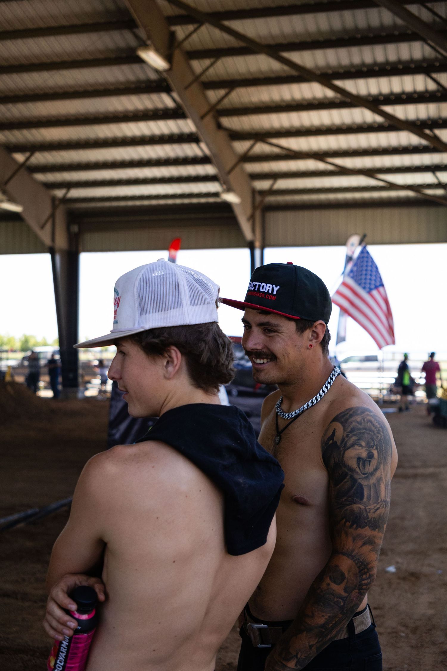 Two shirtless young men with caps and tattoos socialize at an indoor motocross event with US flag.