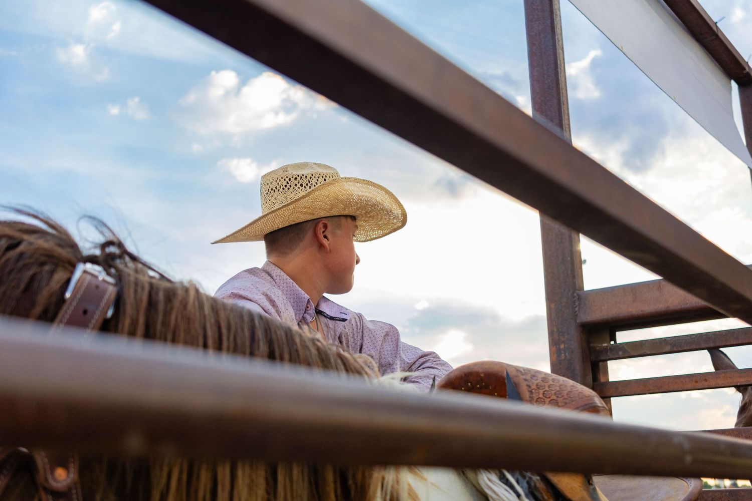 Cowboy in straw hat sits on fence watching rodeo events under a dramatic blue sky at sunset.