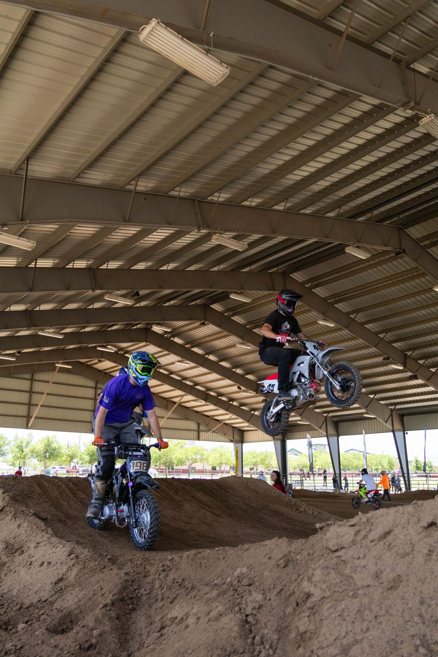 Two motocross riders perform jumps on dirt bikes inside an indoor motocross track with a metal roof.