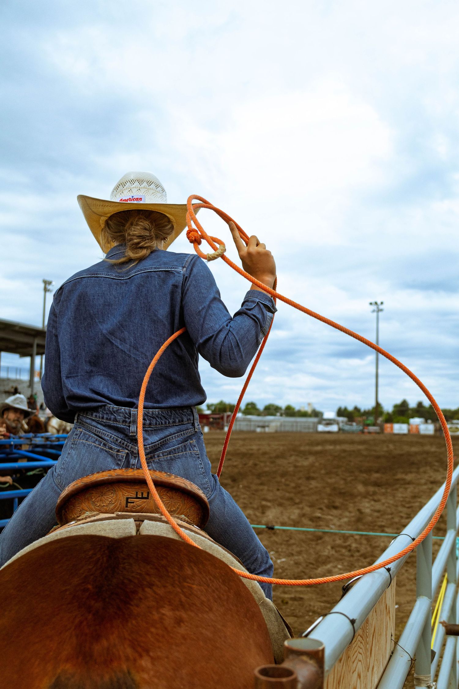 A cowgirl on horseback swings an orange lasso at a rodeo arena, wearing a straw hat and denim jacket.