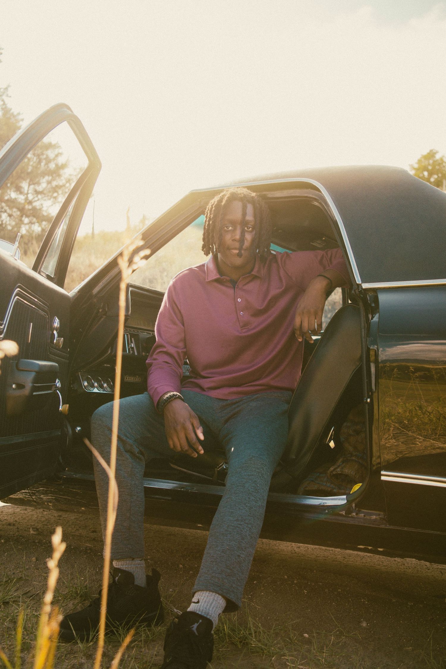 Young man with dreadlocks sitting in the open door of a vintage black car at golden sunset.