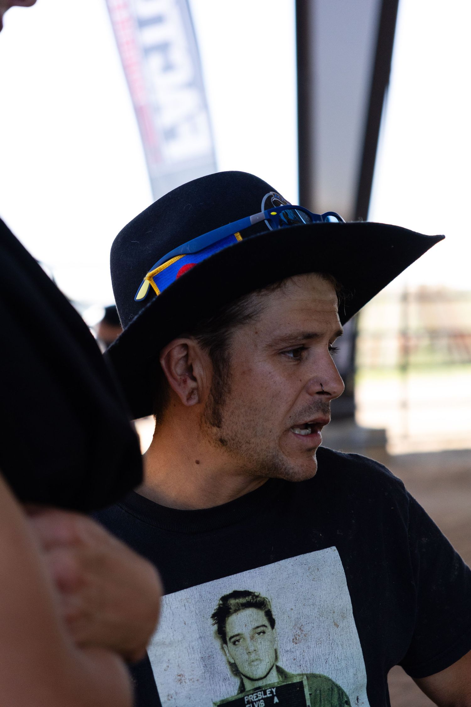 Man wearing a black wide-brim hat with Colorado flag ribbon speaking at an outdoor motocross event.