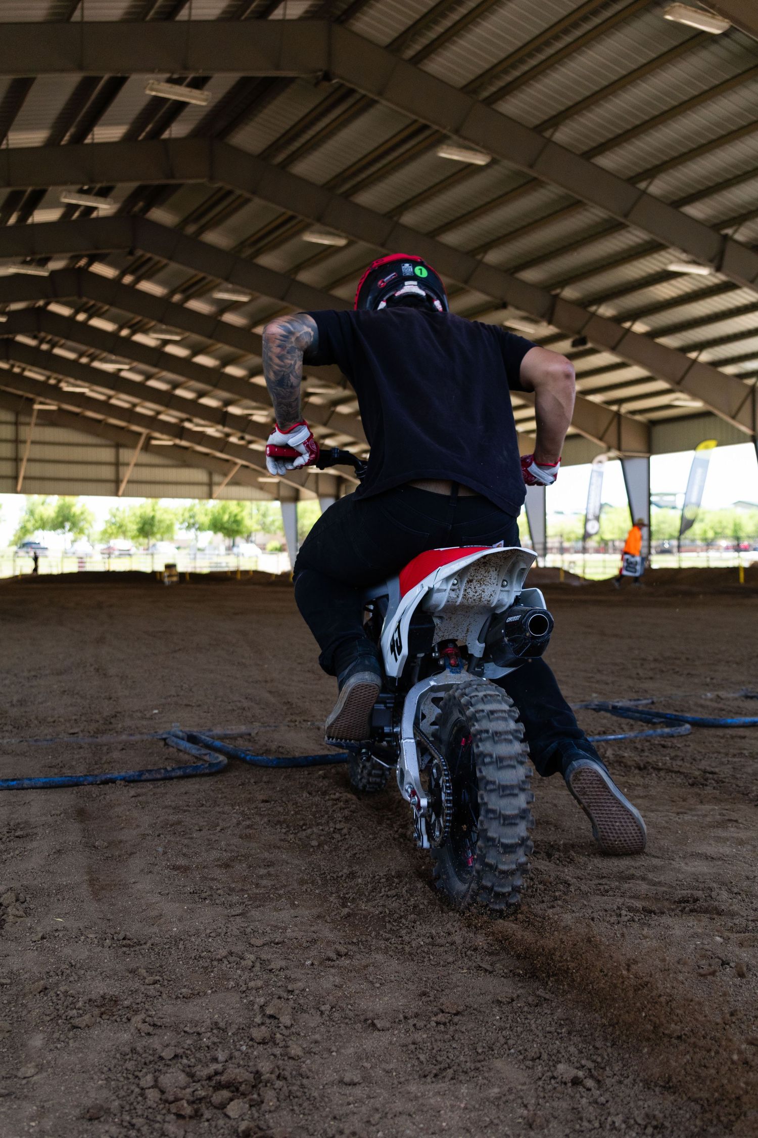 Motocross rider performing a wheelie on a dirt bike inside a covered indoor dirt track arena.