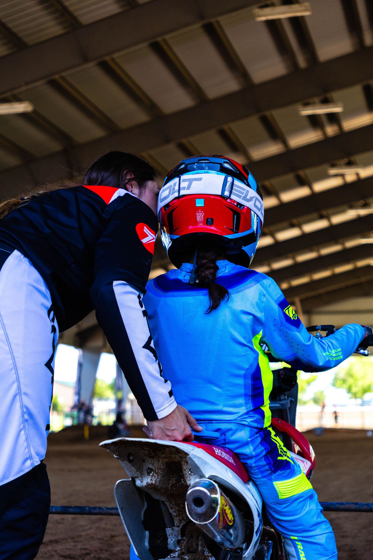 Adult coach assisting a young motocross rider on a dirt bike under a covered track.