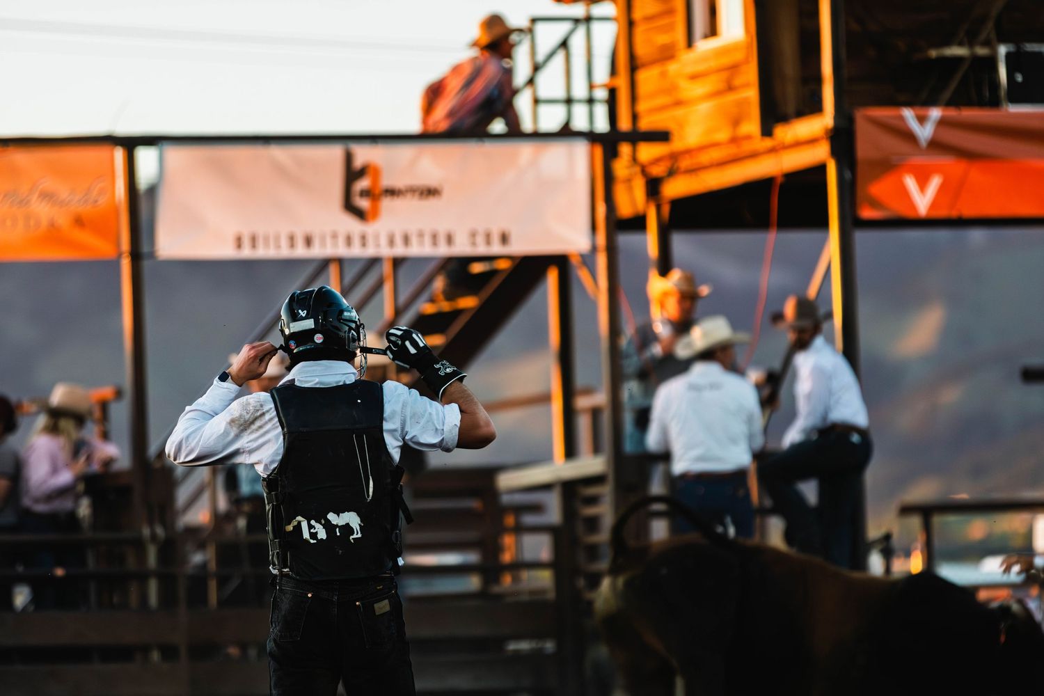 Cameraman filming at an outdoor rodeo event with orange branded signage and cowboys in background.