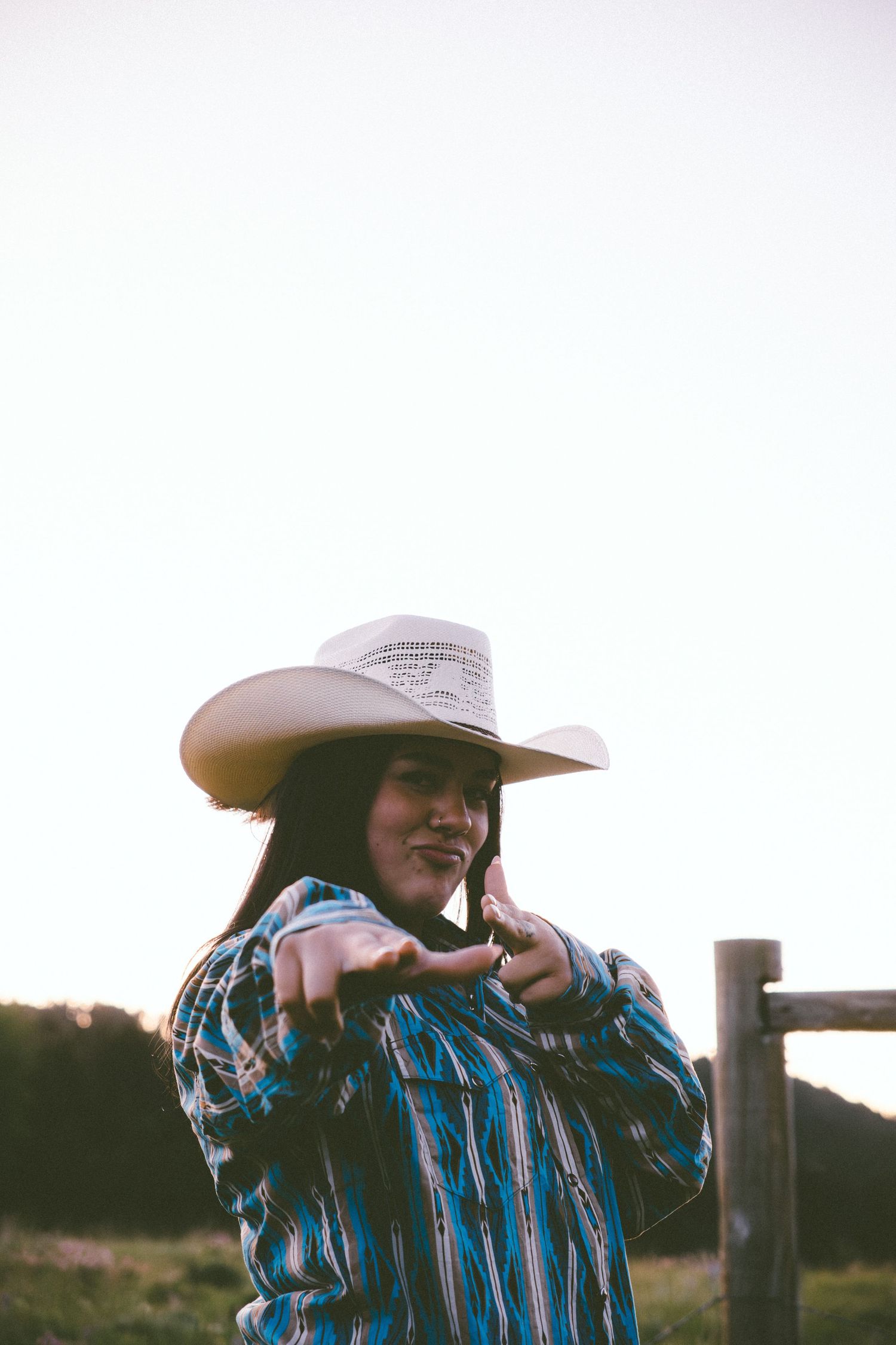 Young woman in cowboy hat and plaid western shirt posing outdoors at golden hour near a wooden fence.