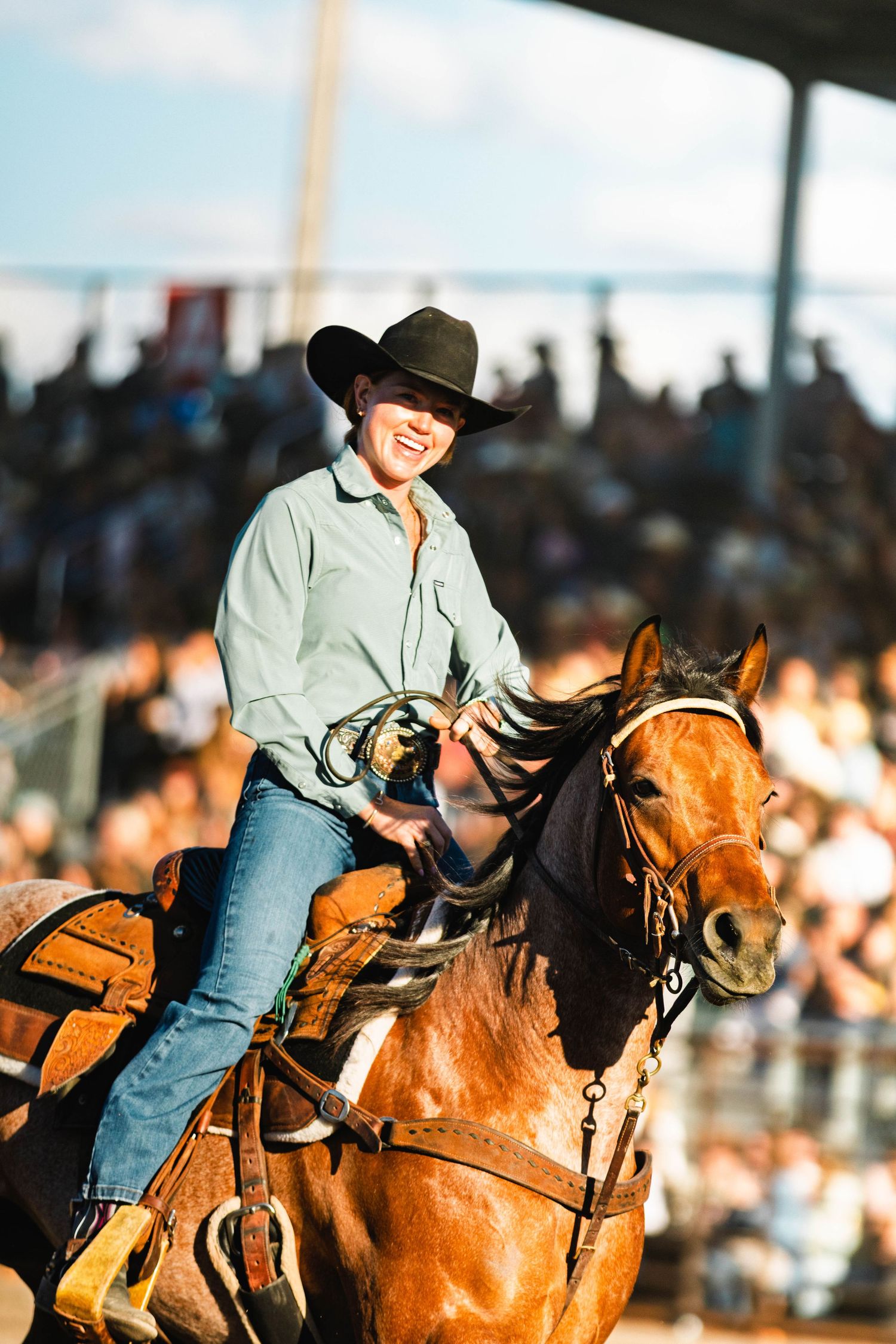 Smiling cowgirl in black hat rides a chestnut horse at a rodeo arena with crowd in background.