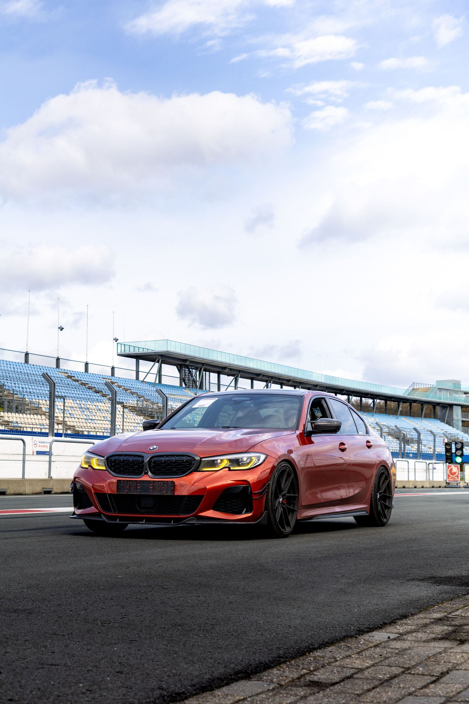Red BMW M3 sedan parked on a racing circuit pit lane with grandstands visible in the background.