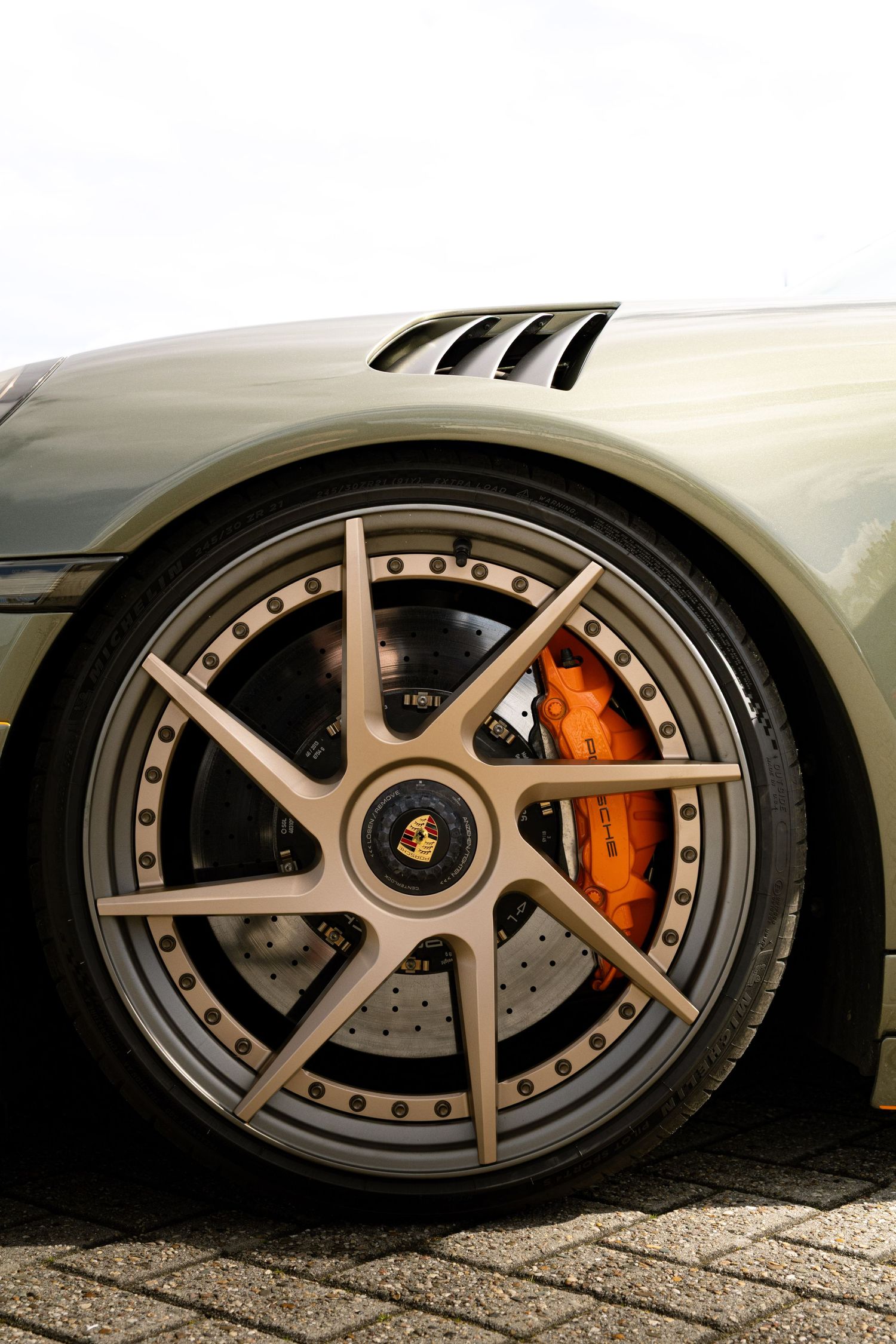 Close-up of a Porsche sports car wheel featuring a gold multi-spoke rim and bright orange brake calipers on cobblestone.