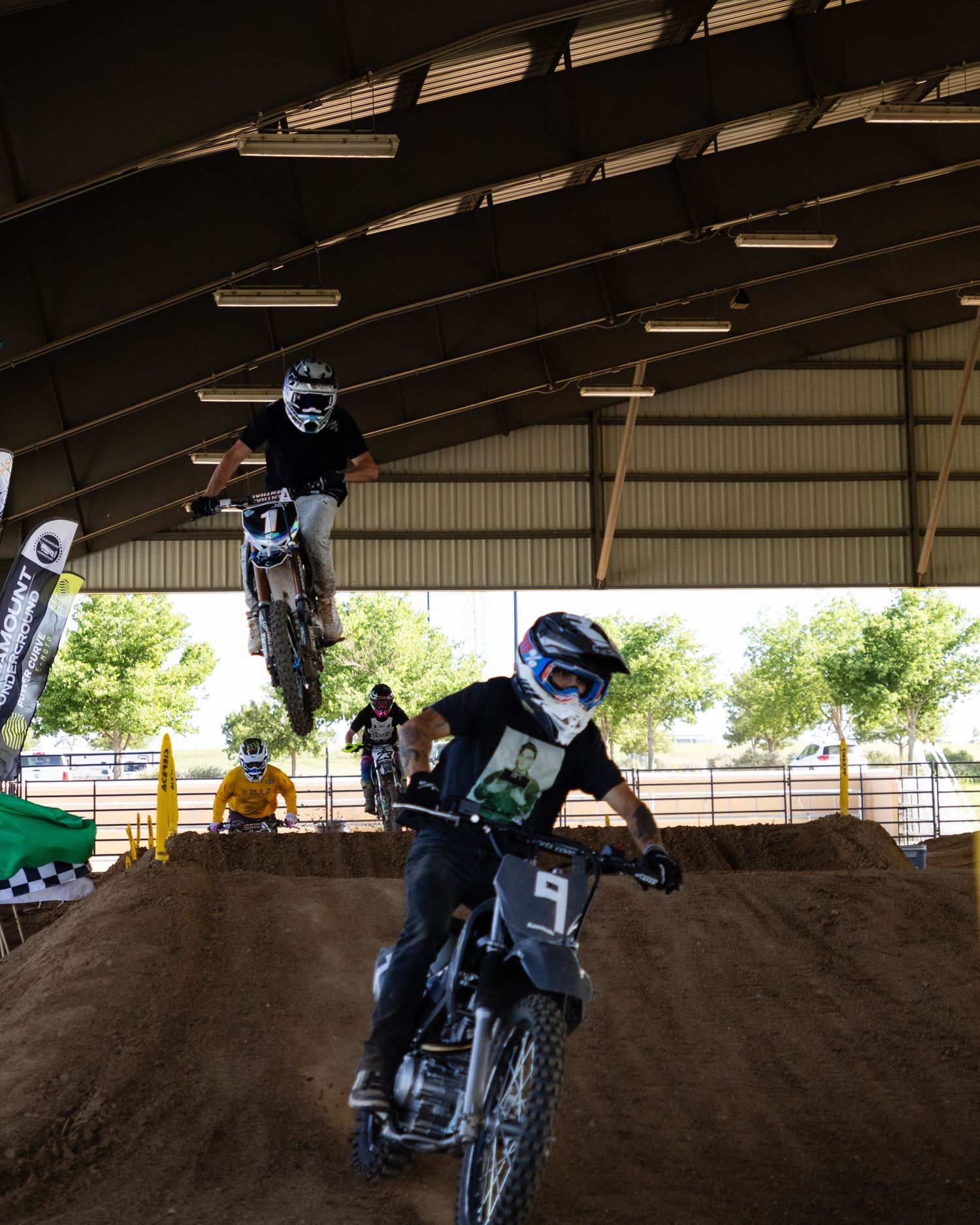 Two motocross riders race on indoor dirt track, one catching air while other navigates berm.