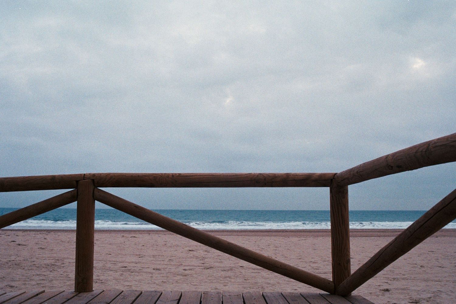 Wooden beach boardwalk railing overlooking a sandy shore and overcast ocean horizon.