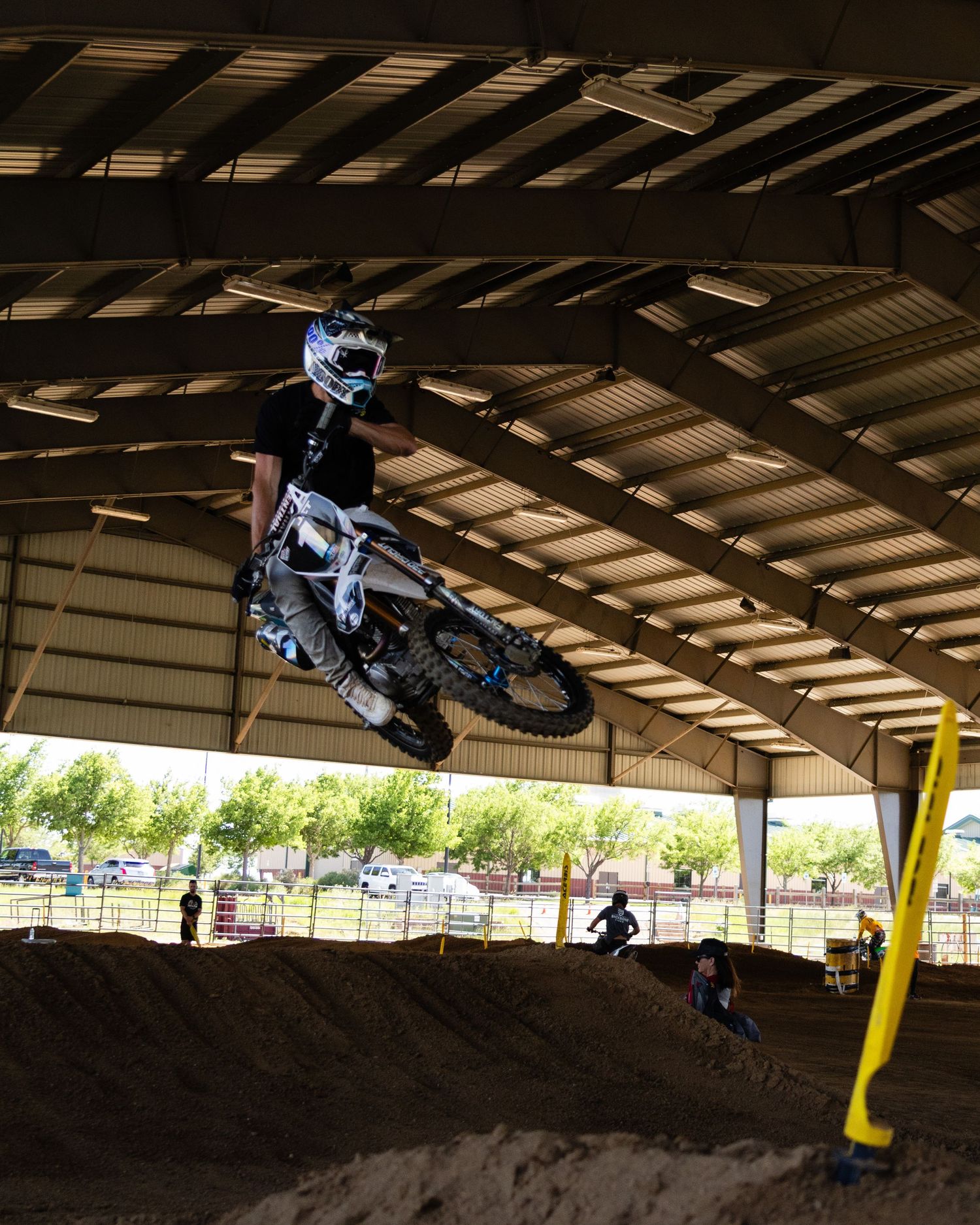 Motocross rider in black and white gear jumps high over dirt berm at indoor arena.