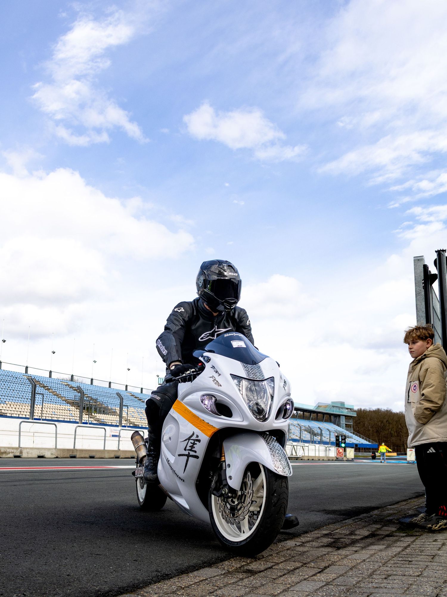 Motorcyclist in full gear rides a white Suzuki Hayabusa on a racing circuit under a cloudy sky.