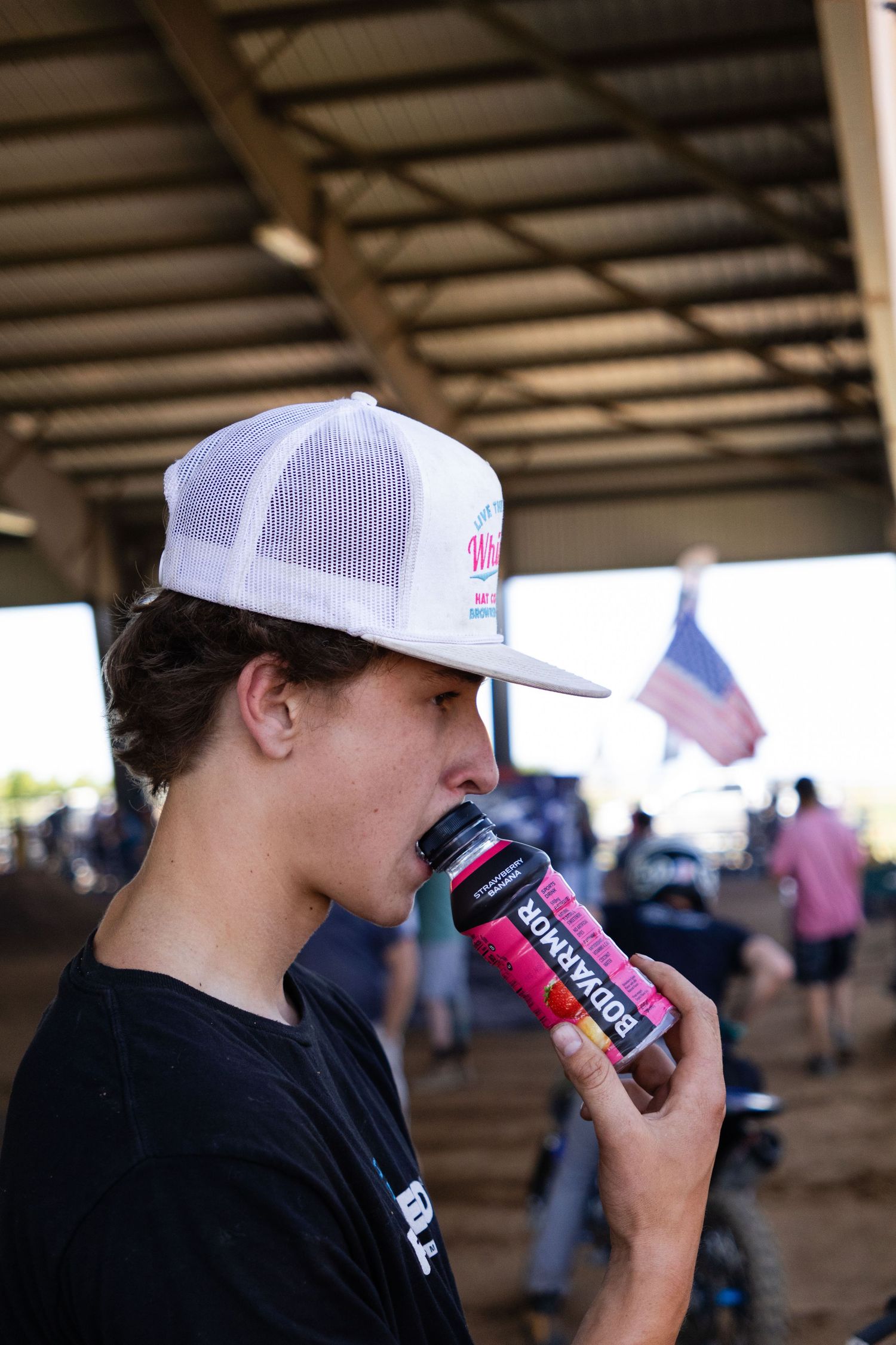 Young man in white snapback cap drinking a BODYARMOR sports drink at an outdoor motocross event.
