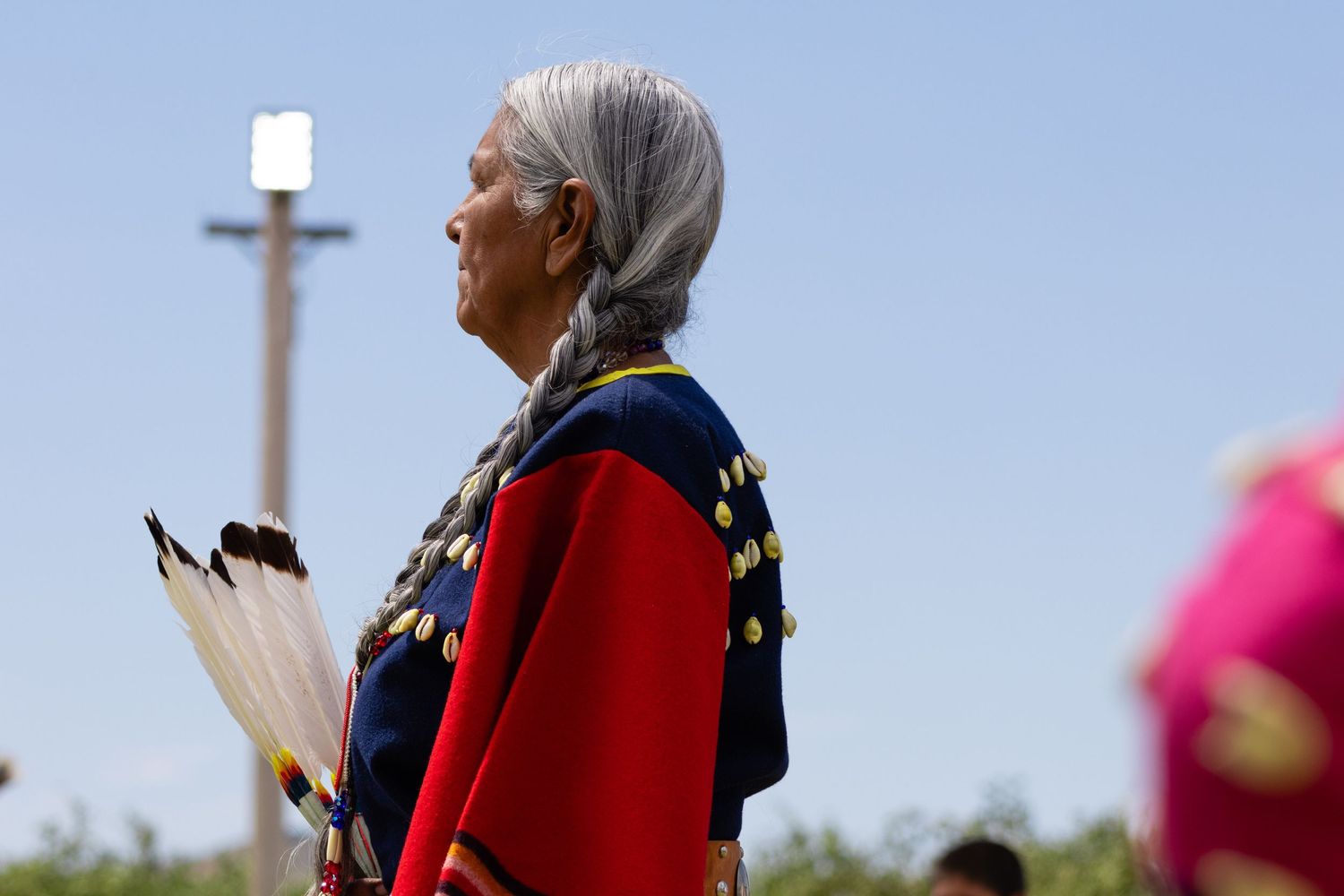 Elderly Native American woman in red and blue traditional dress holding feathers at an outdoor powwow.