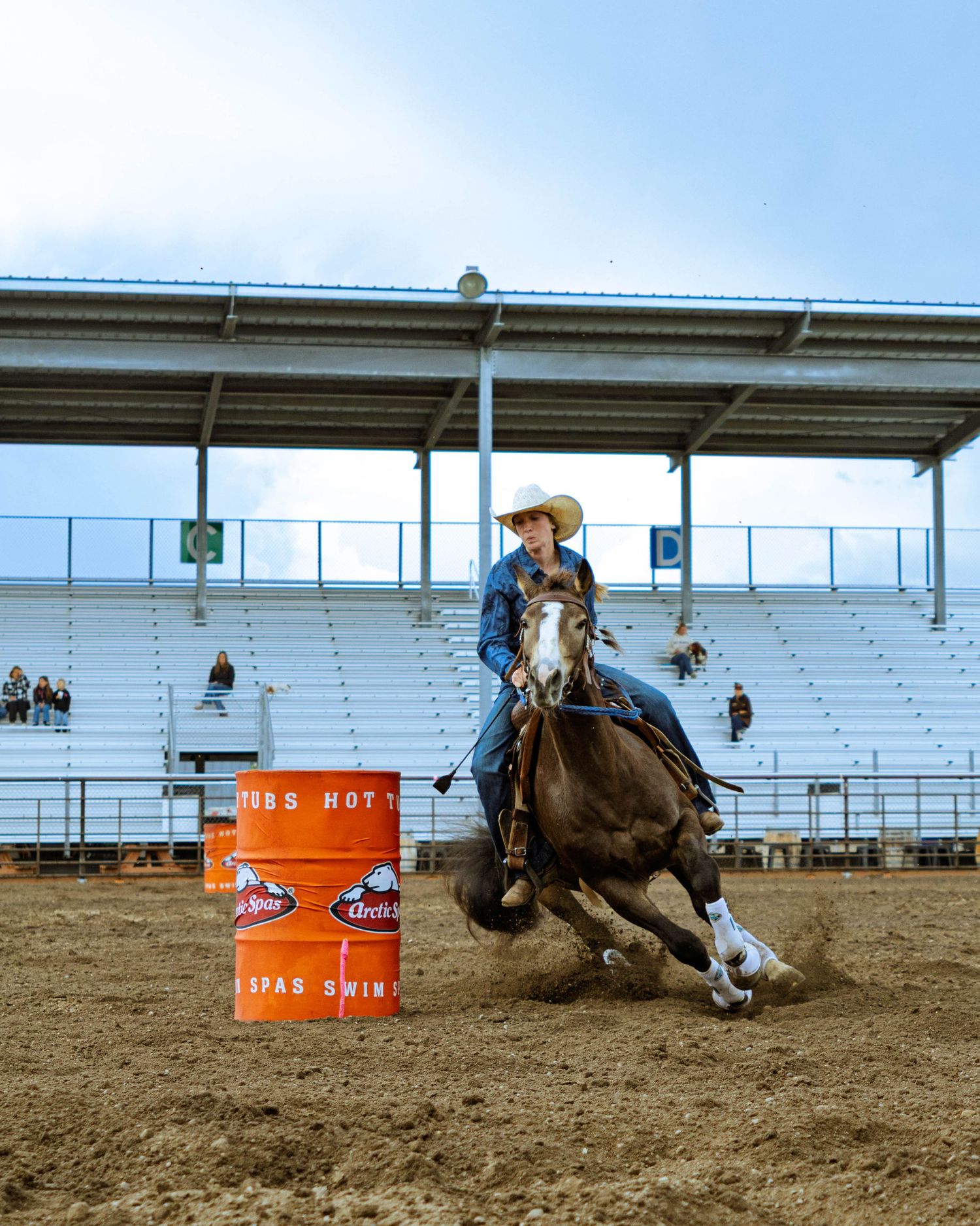 A cowgirl rides a horse around an orange barrel during a rodeo barrel racing competition in a sandy arena.