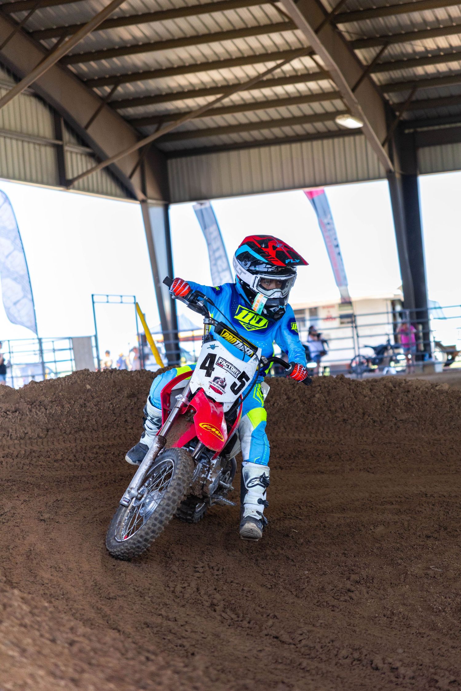 Young motocross racer in blue gear rides a red Honda through an indoor dirt track corner.