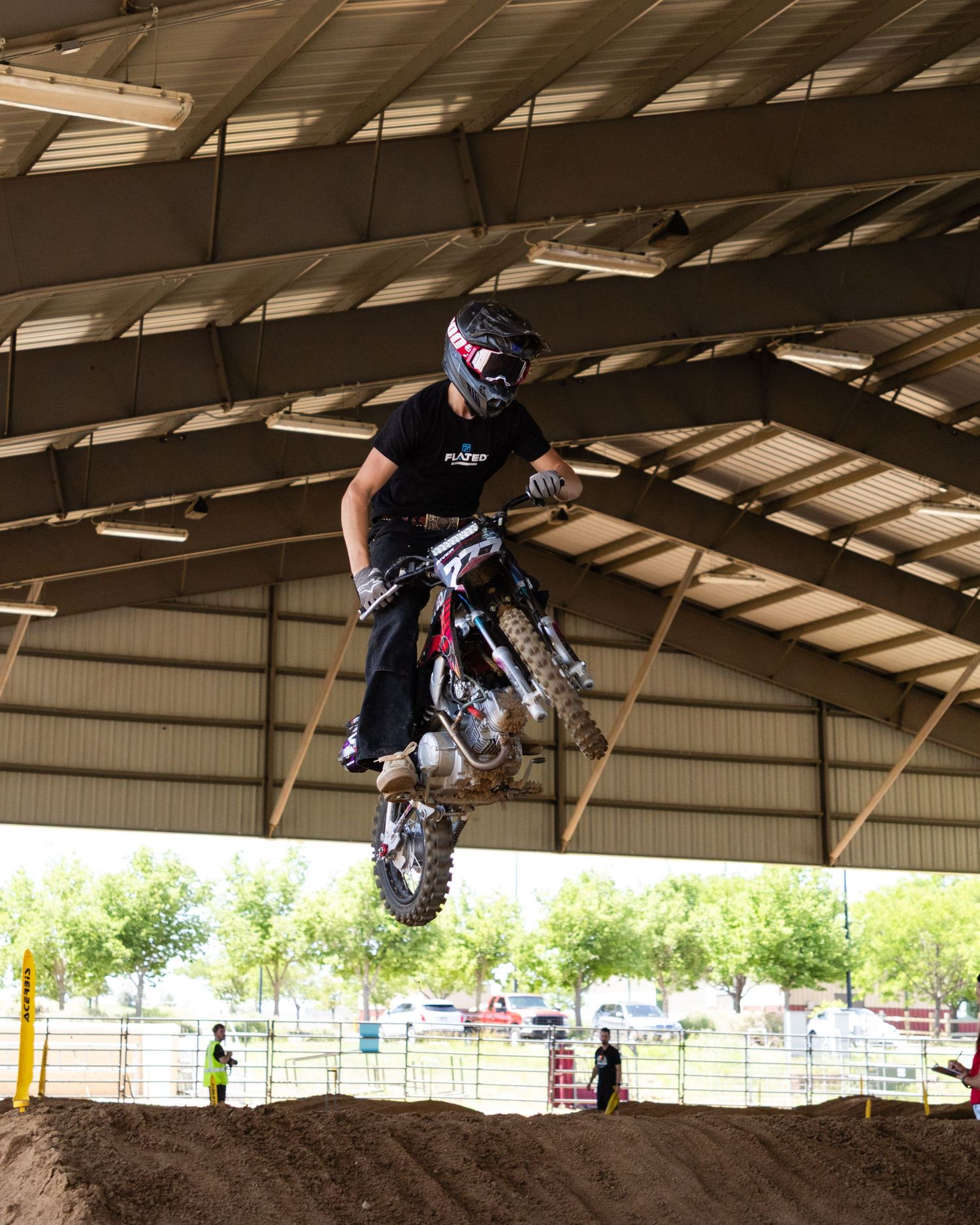 Motocross rider catching air off a dirt jump inside a large covered arena during a supercross competition.