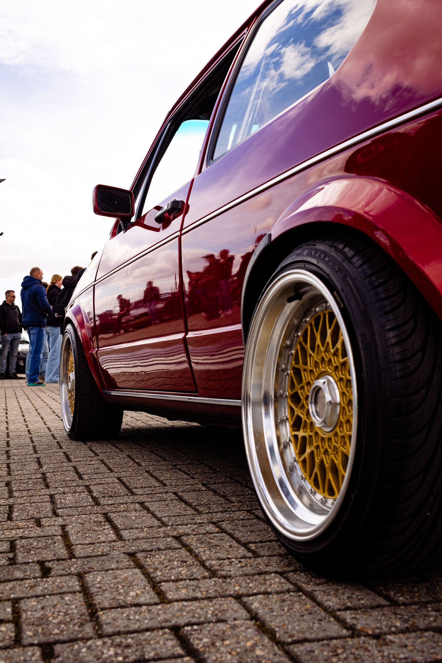 A lowered red car with gold BBS wheels on cobblestone at an outdoor car show.