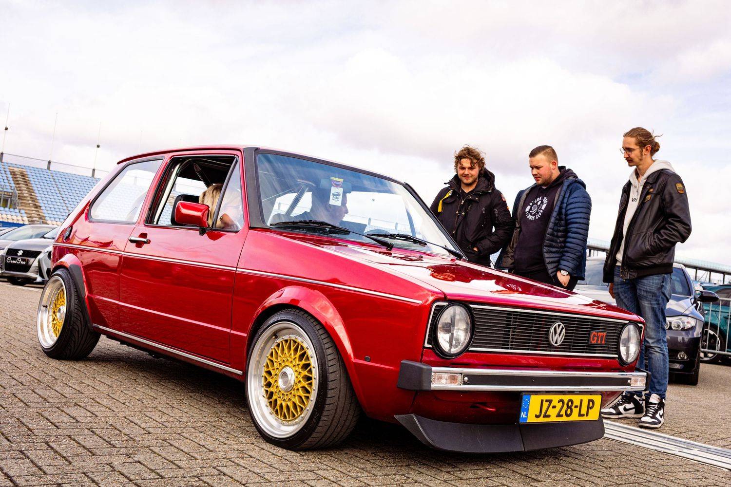 Three people admiring a red Volkswagen Golf Mk1 with gold BBS wheels at a car meet in the Netherlands.