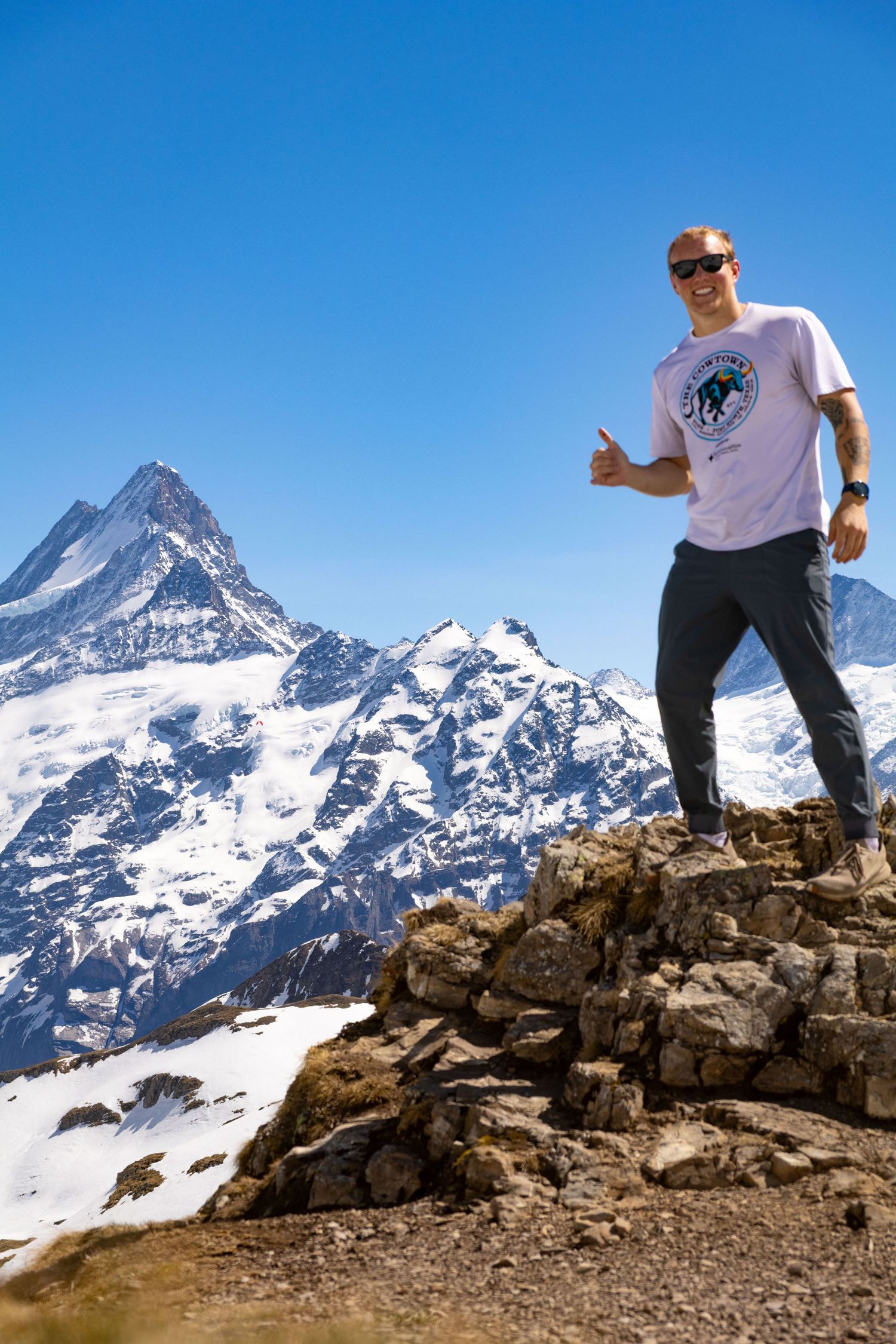 Happy male hiker giving thumbs up on rocky summit with snow-capped mountain peaks behind him.