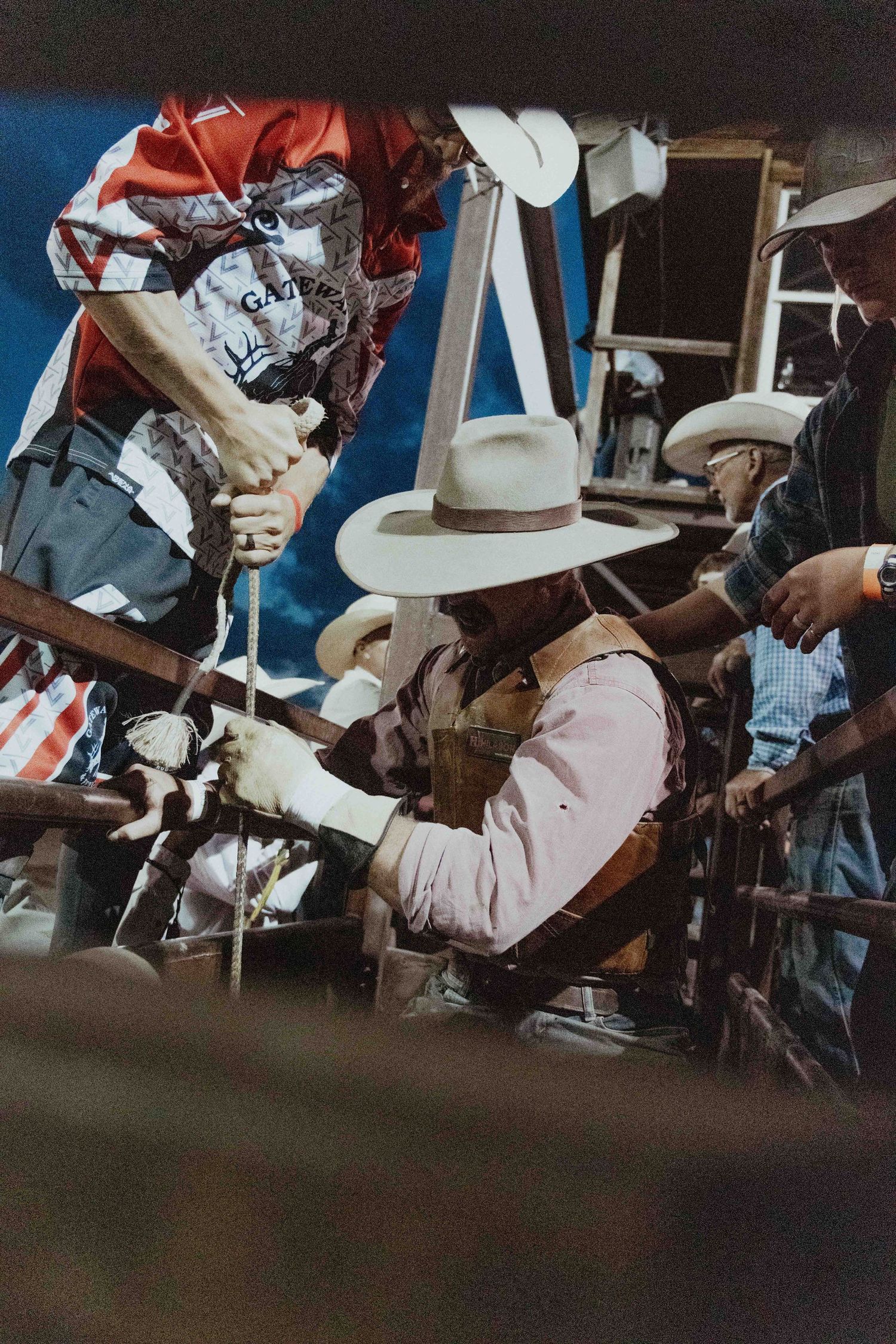 Cowboys in hats preparing a bull rider in the chute before a rodeo competition at night.