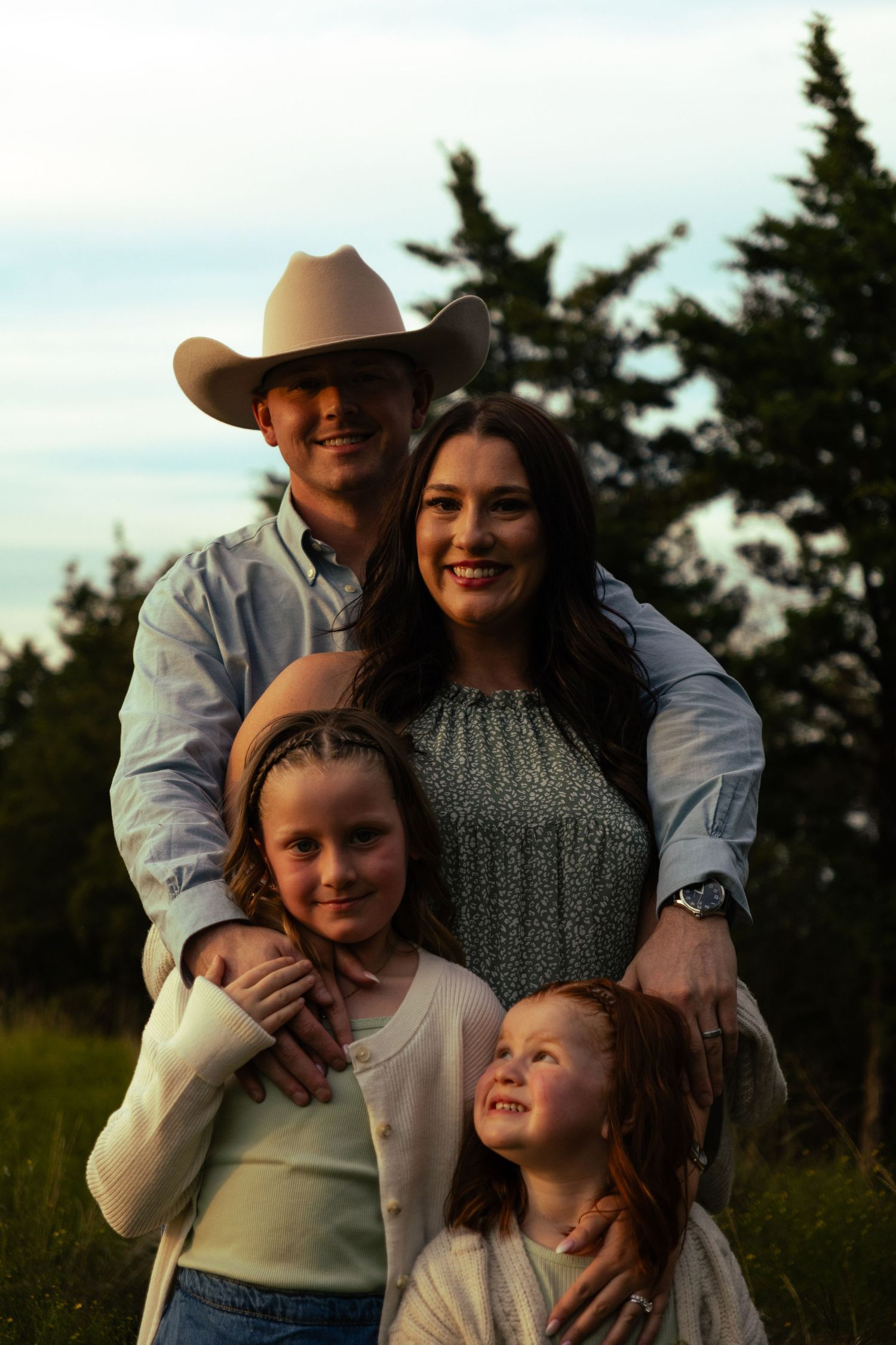 Smiling family of four at sunset in a grassy field, father in cowboy hat with mother and two daughters.